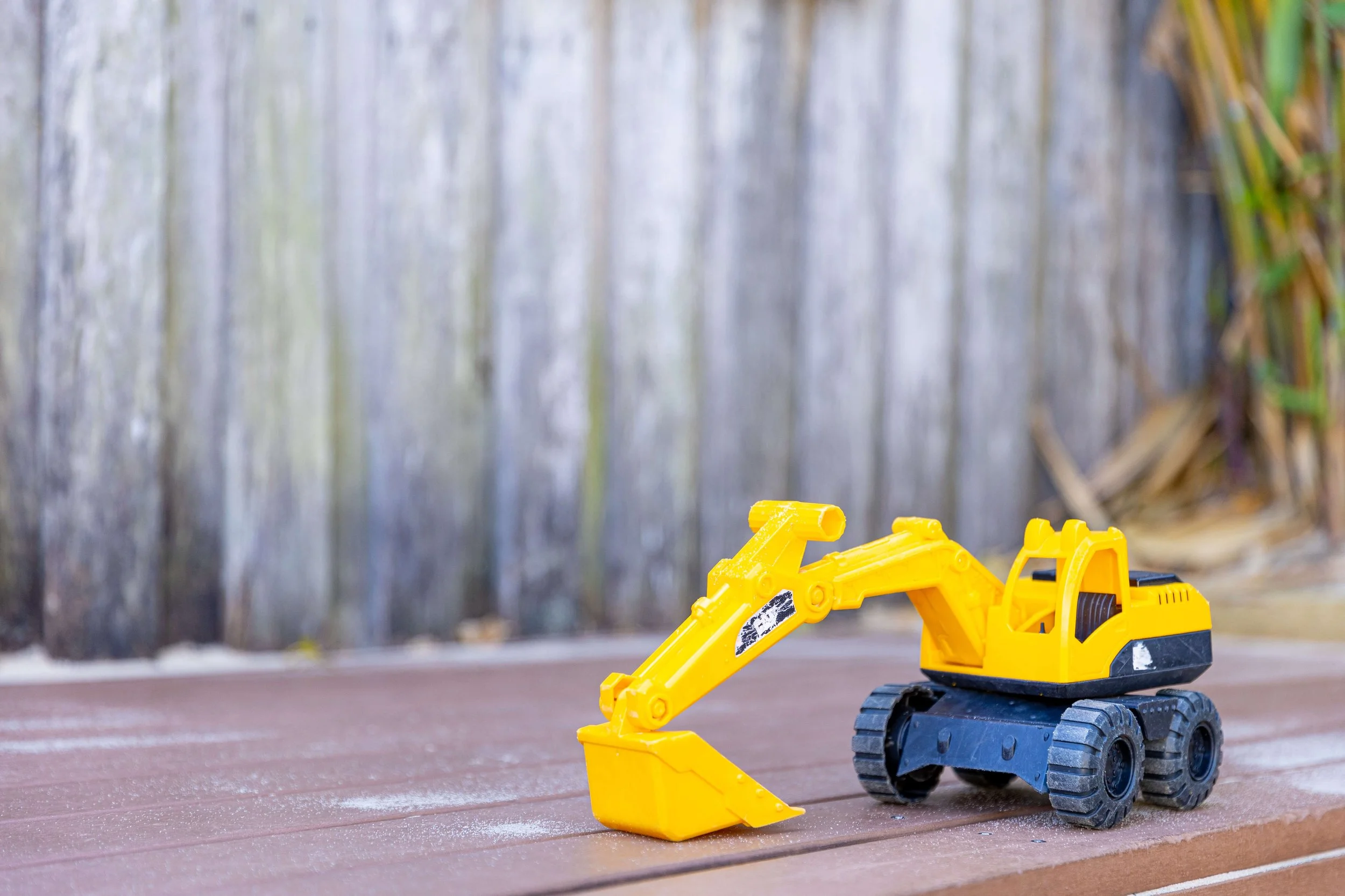 A yellow toy excavator on a wooden deck with a wooden fence and some plants in the background.