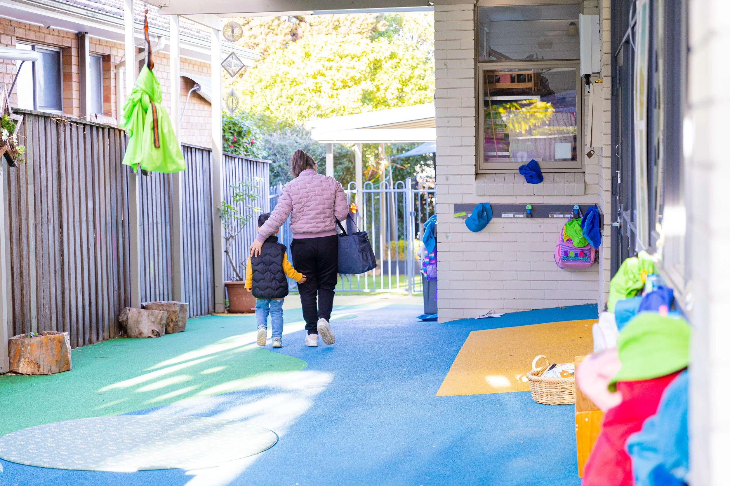 A woman and child walking toward a gate outside a preschool, with colorful outdoor play area and backpacks on hooks.