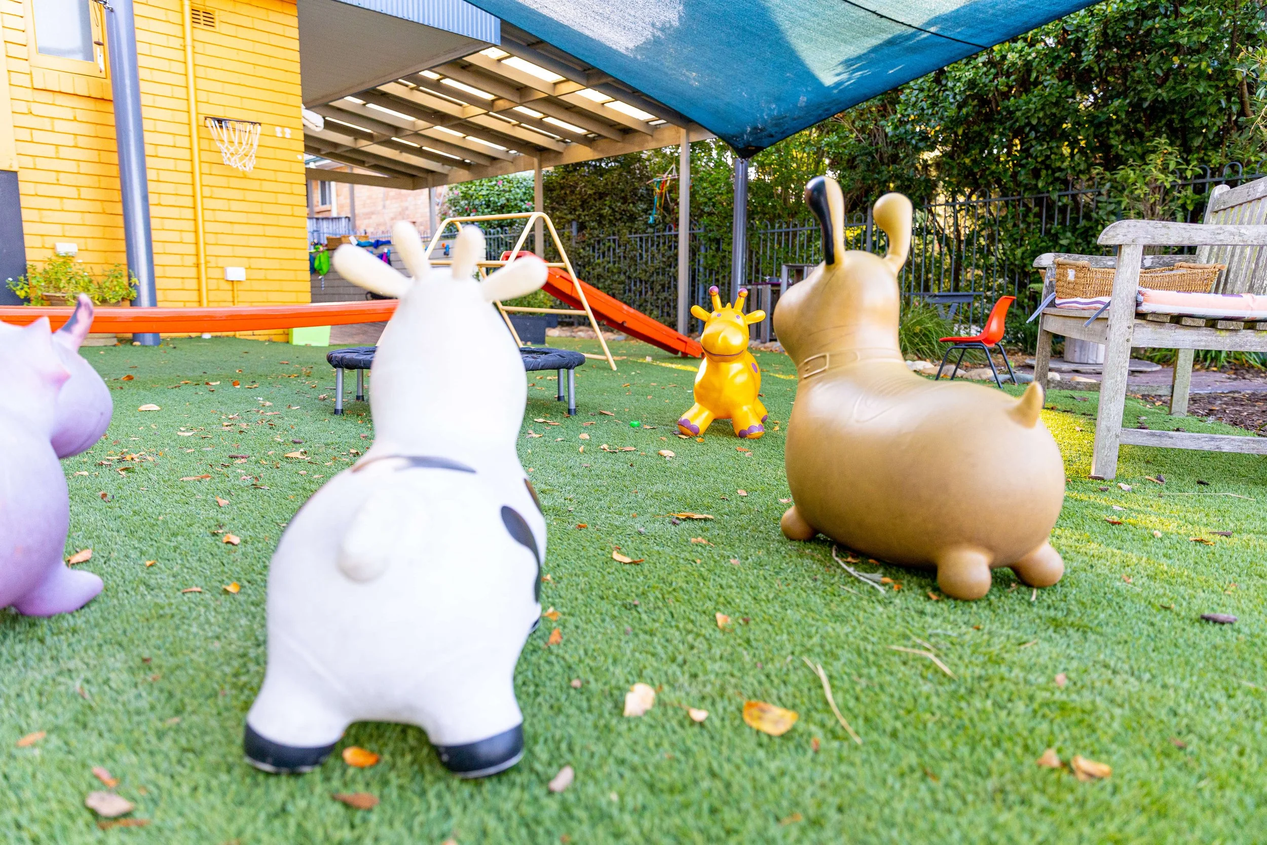 Colorful outdoor children’s play area with animal-shaped playground toys, including a white cow, a brown dog, and a yellow giraffe, on green artificial turf with a slide in the background, shaded by a blue canopy.