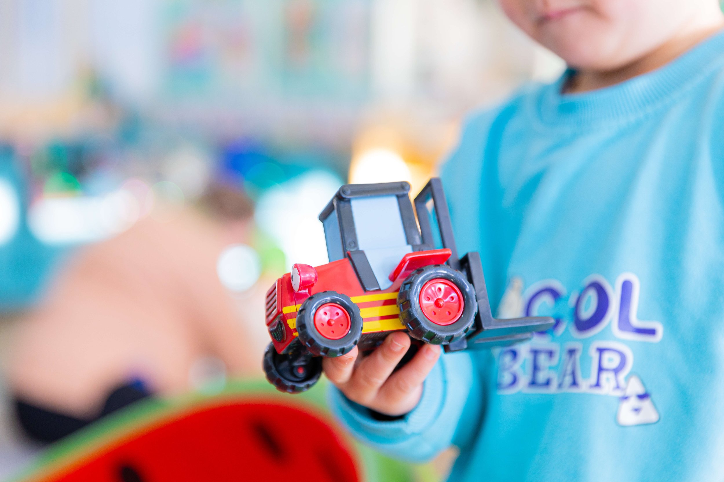 Child holding a red toy tractor in a colorful, blurred background.