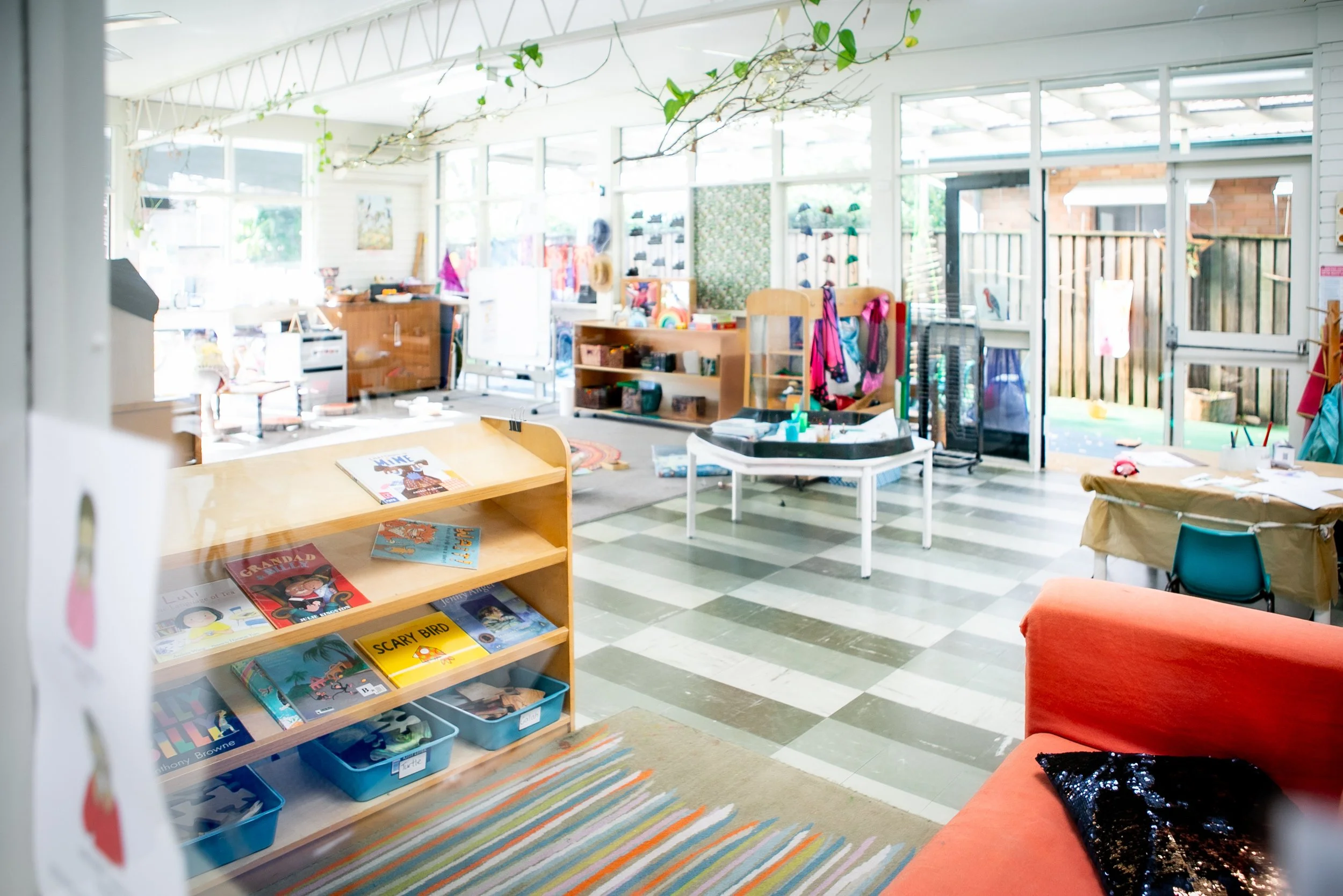 Bright, colorful classroom or playroom with bookshelves, tables, chairs, and toys. Large windows and a glass door let in natural light, and there is a plant hanging from the ceiling.