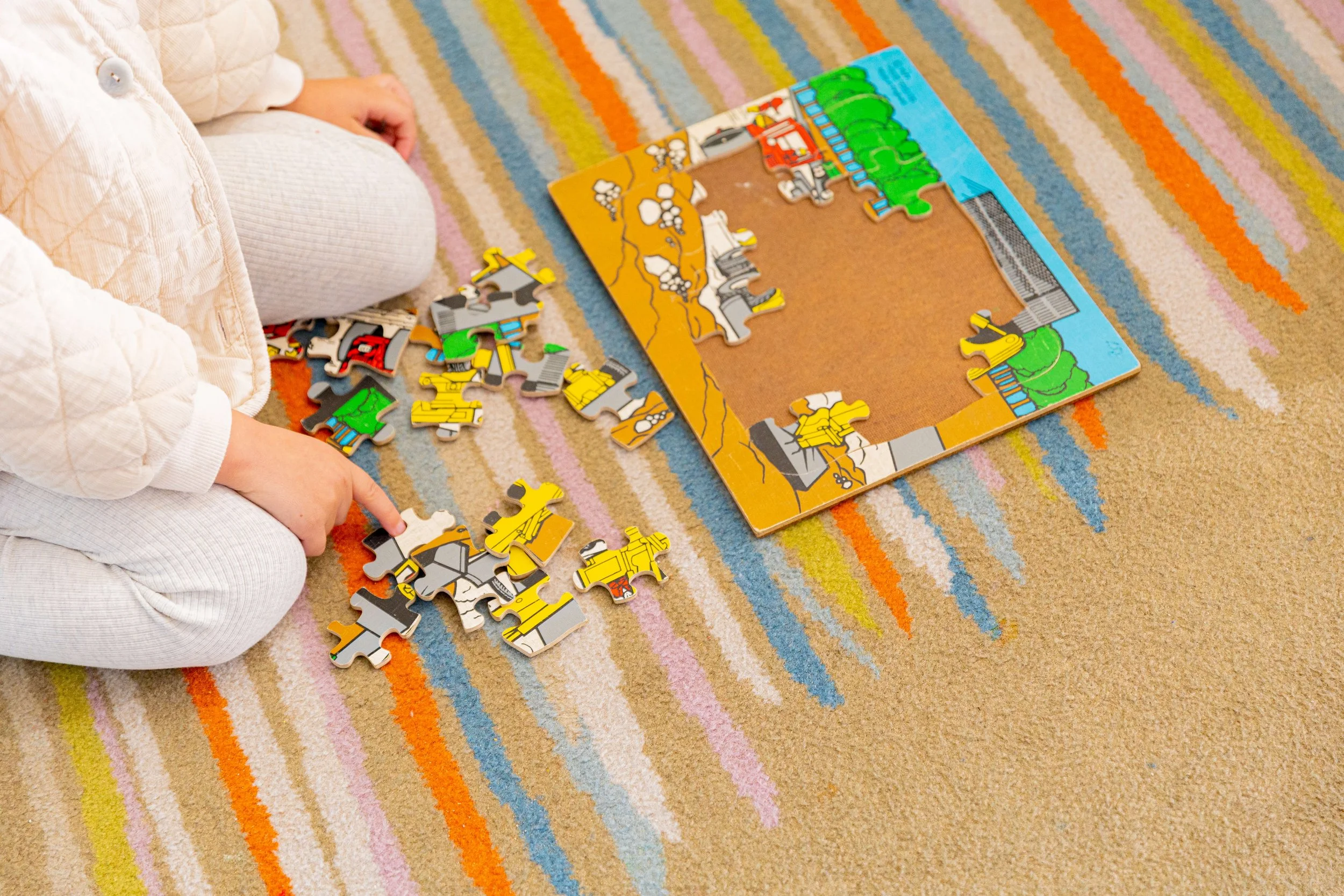 A child sitting on a colorful striped carpet, assembling a puzzle that depicts a construction scene.
