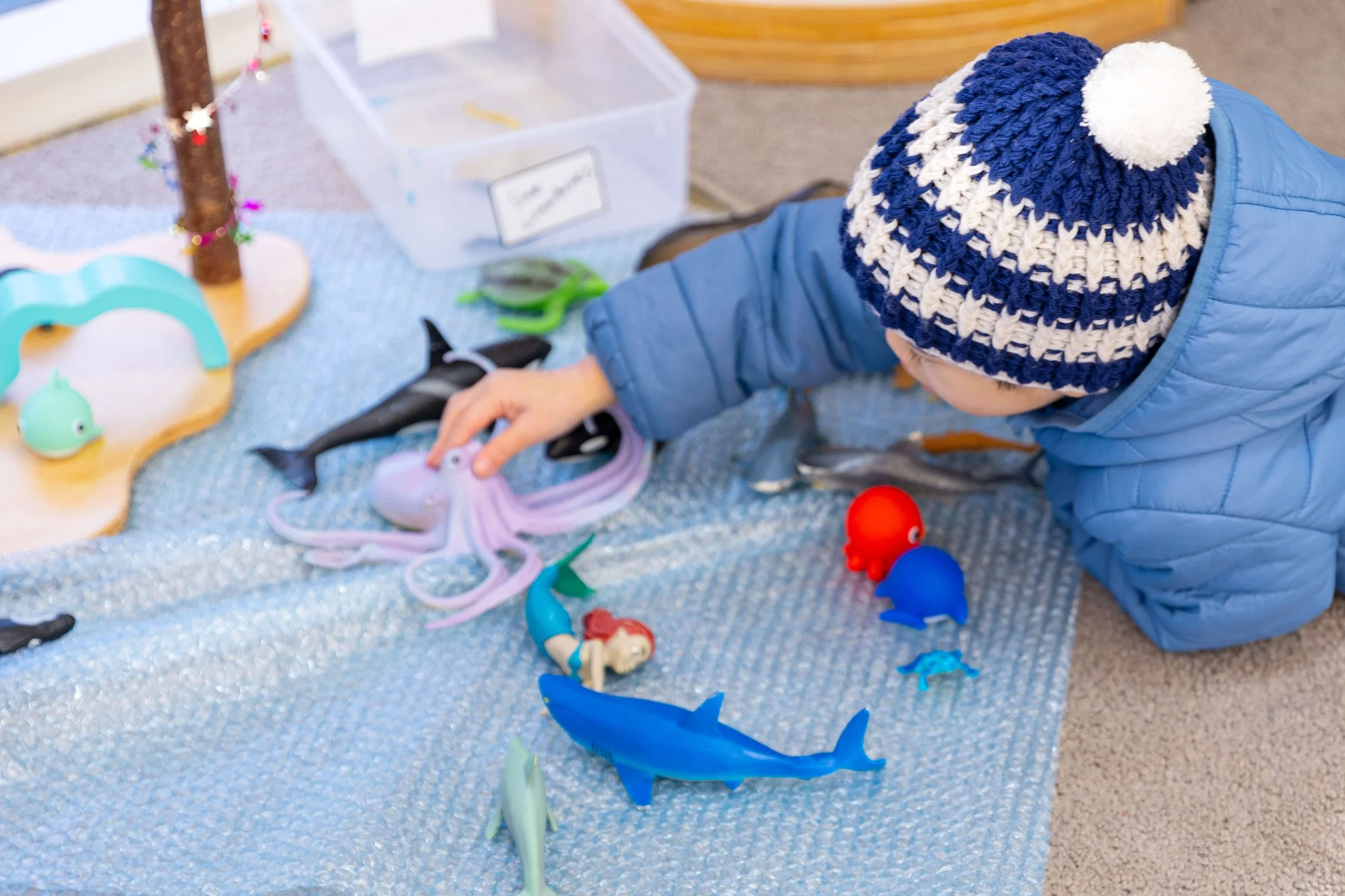 Child wearing a blue jacket and a blue and white knit winter hat playing with toy sea creatures on a blue textured mat.