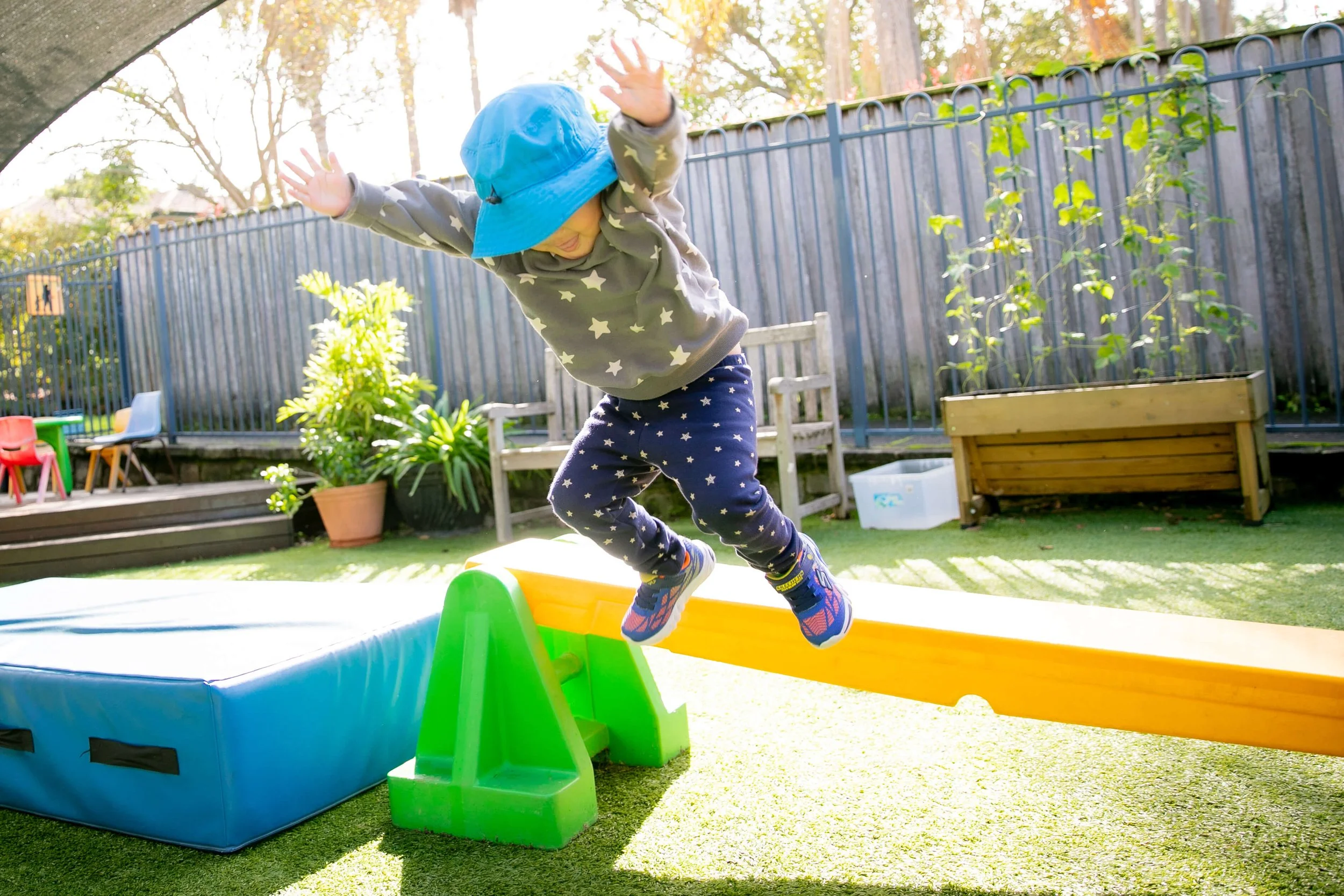 Child wearing a gray sweatshirt with star patterns, navy star leggings, and a blue hat jumps off a yellow balance beam in a backyard with toys, plants, and a wooden fence.