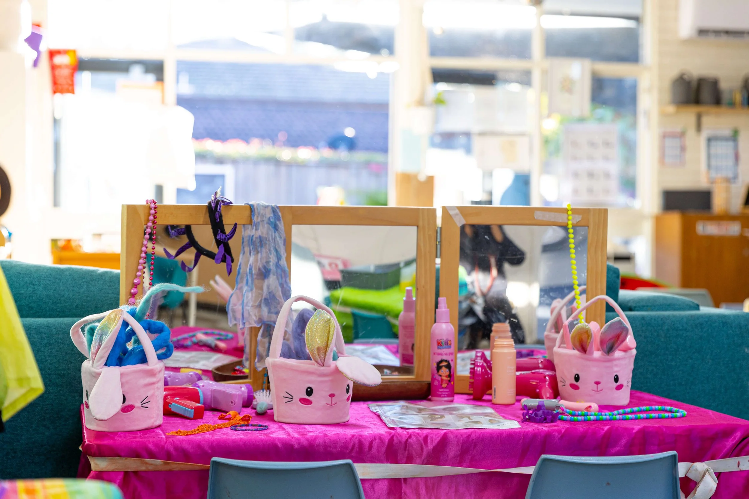 A colorful table with pink tablecloth displays children's toys, accessories, and makeup items, including bunny-themed bags, bottles, and a mirror, set against a bright, windowed room.