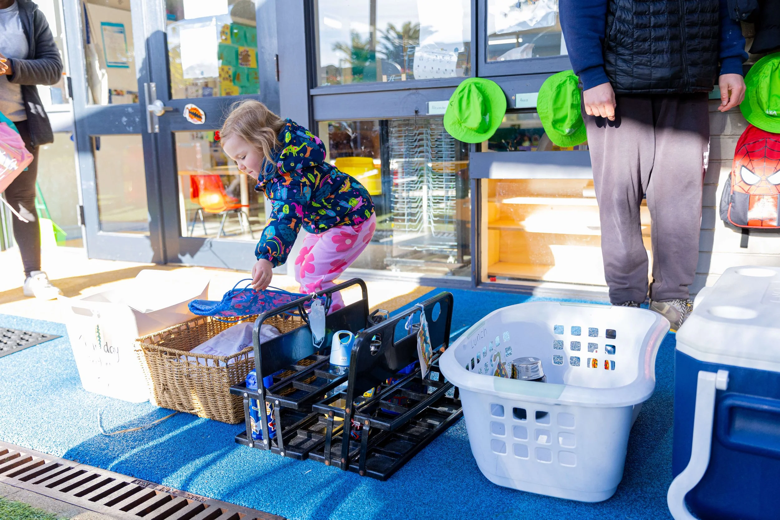 A young girl with blonde hair, wearing a dark blue jacket with colorful patterns and pink pants with large flowers, is picking up a blue object from a basket outside a building with a glass door and windows. An adult male, wearing gray pants and a bl