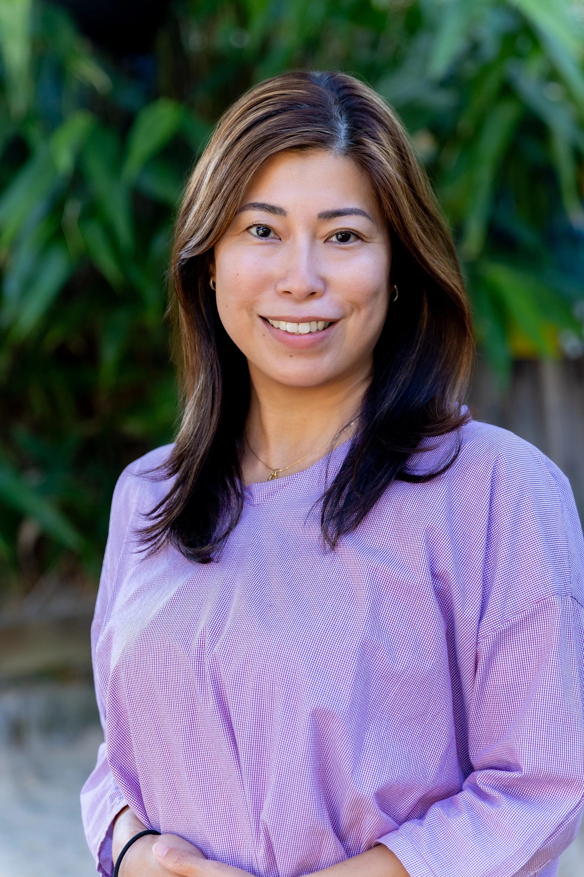 Portrait of a smiling woman with long dark hair, wearing a pink sweater, standing outdoors with colorful blurred background.