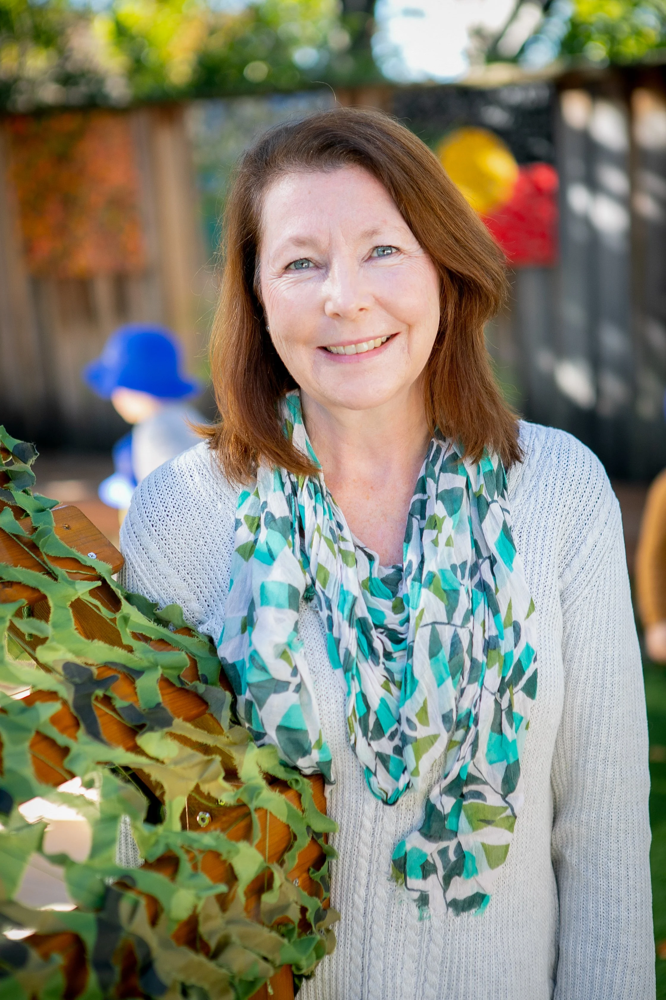 A woman with light skin and brown hair smiling outdoors, wearing a white sweater and a patterned scarf, with a child in a blue hat in the blurred background.