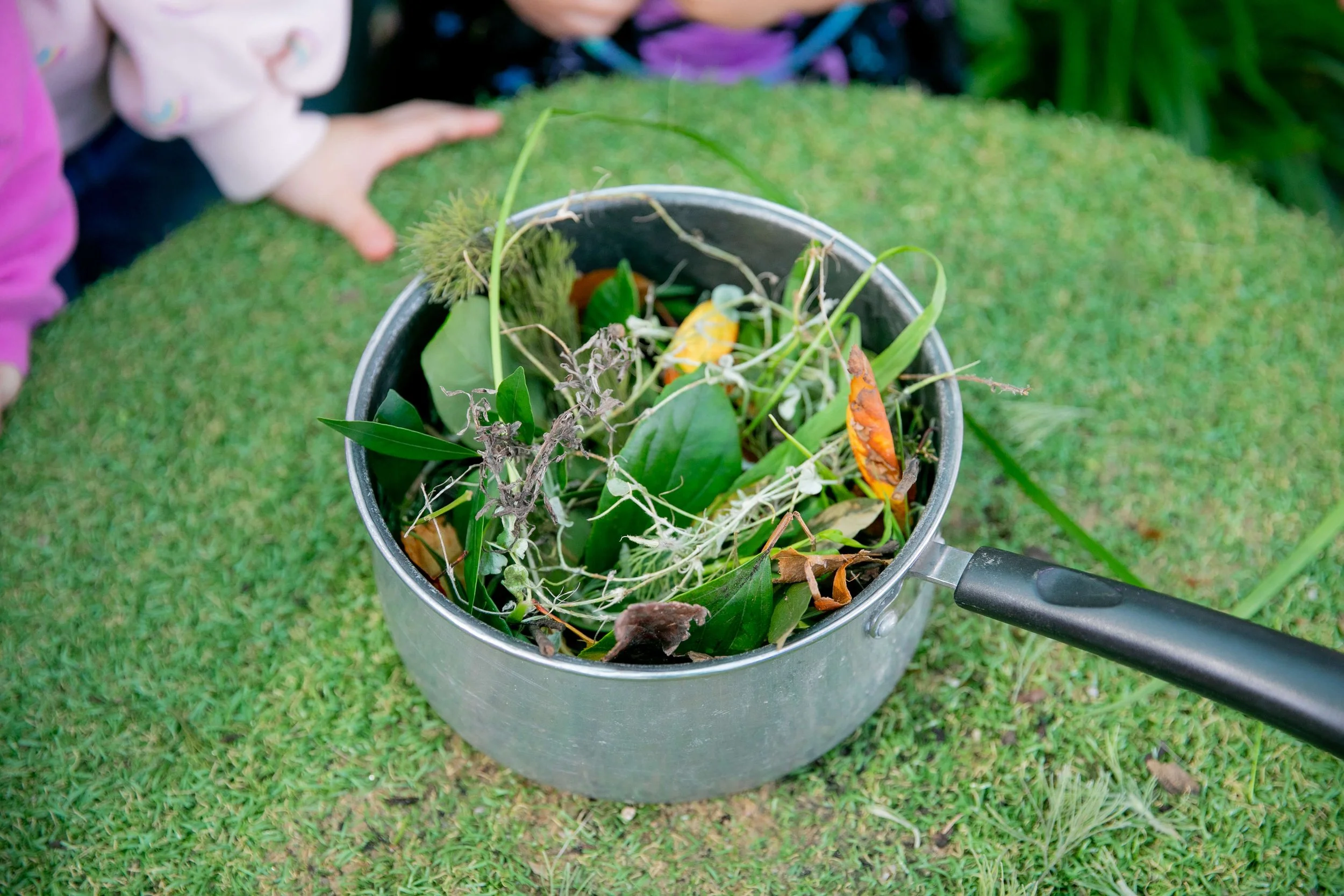 A metal pot filled with dead leaves, twigs, and plant debris on a grassy surface, with children nearby.