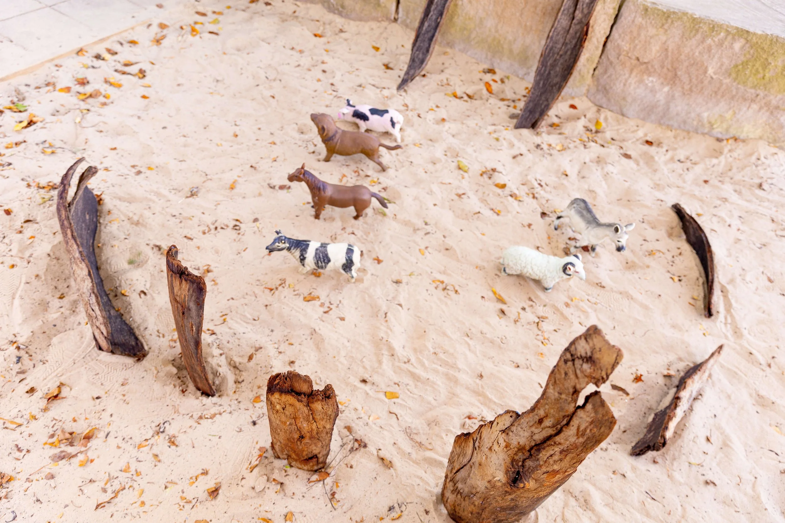 Miniature farm animals including cows, sheep, and goats on a sandy surface with wooden logs arranged in a circle.