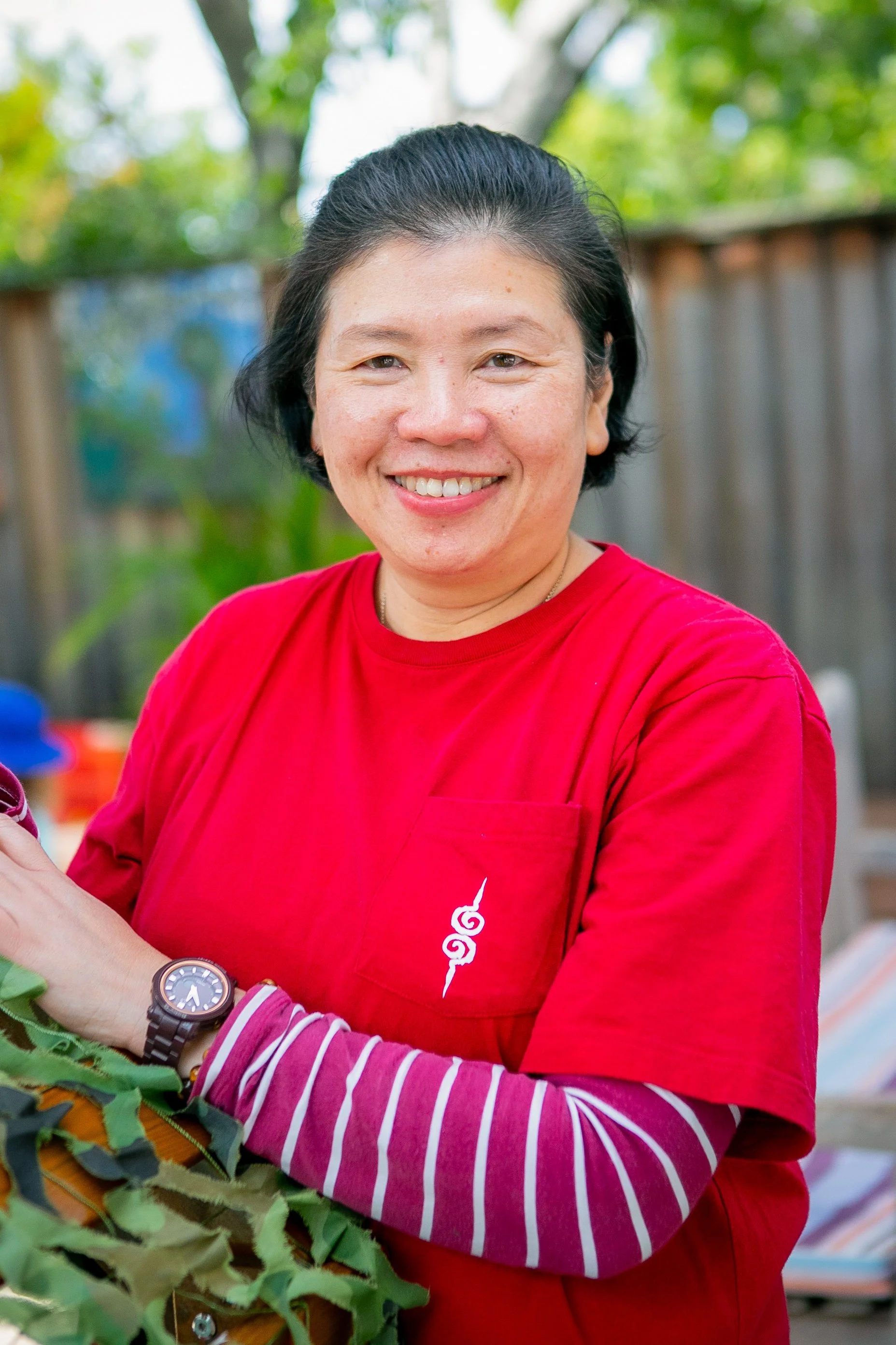 A woman smiling outdoors, wearing a red shirt with a small white design on the pocket. She has short black hair, a watch on her left wrist, and is holding some green leafy foliage. Trees and a wooden fence are in the background.