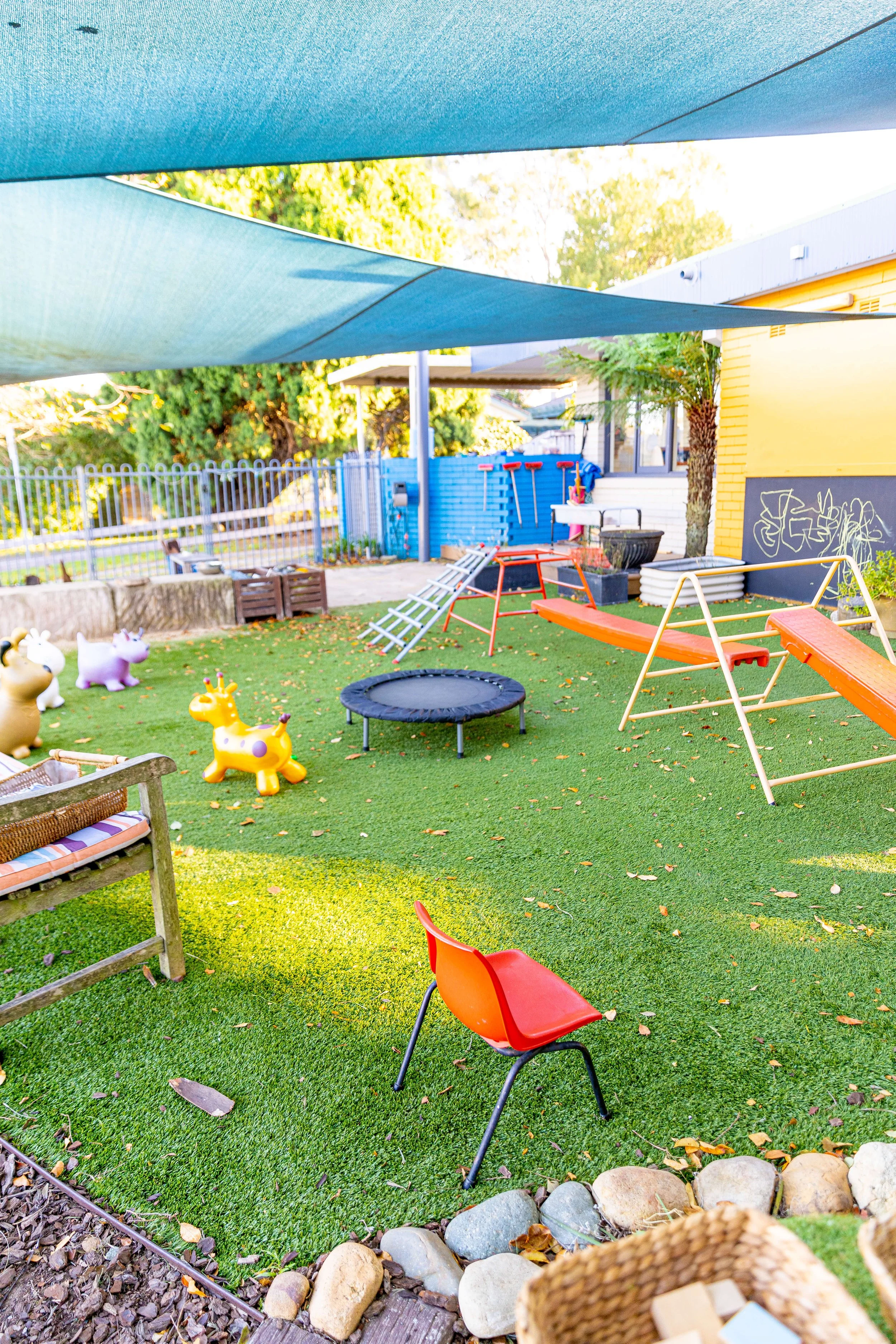Colorful outdoor playground with small slides, a trampoline, riding toys, and chairs on artificial grass.