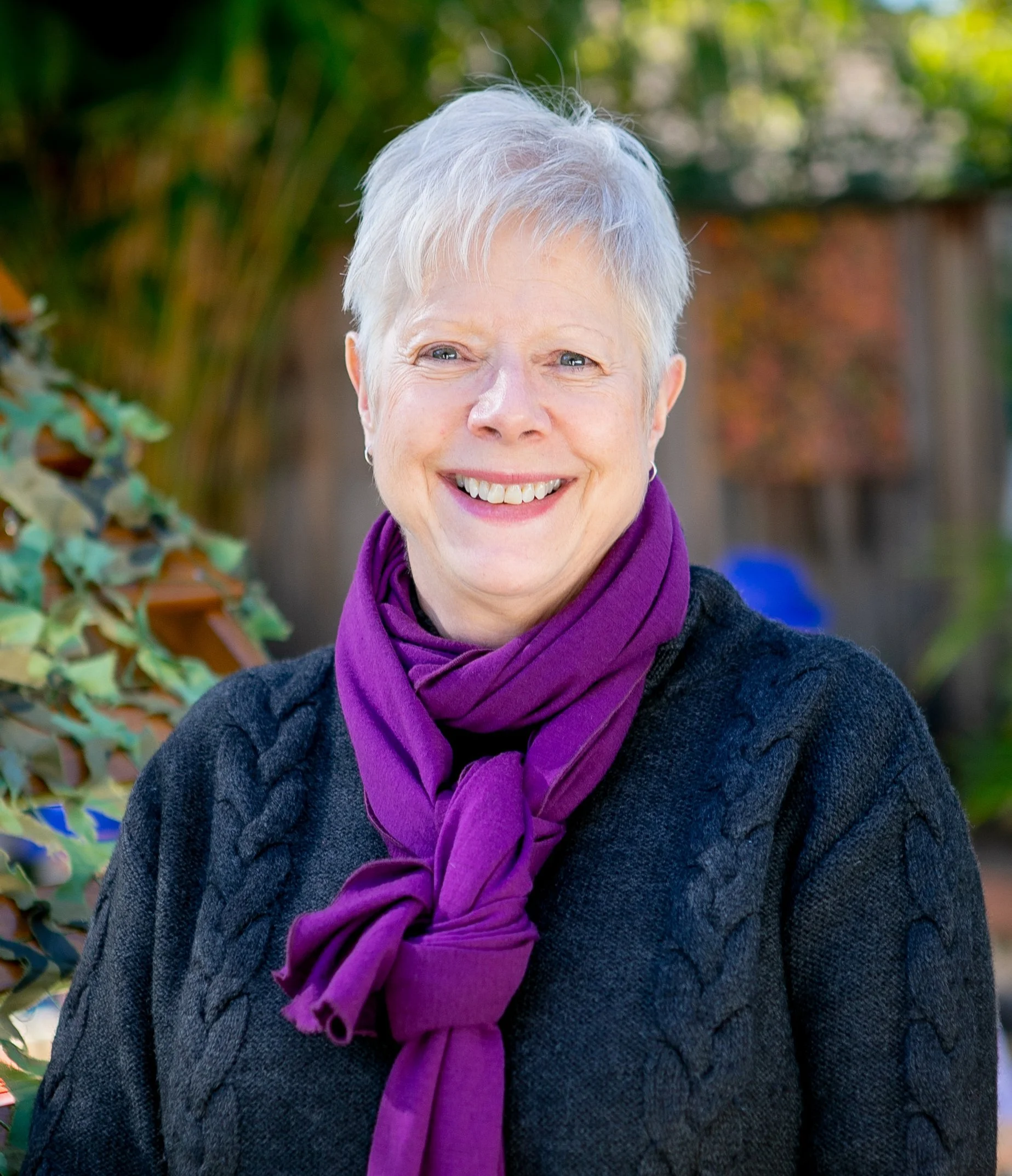 A smiling woman with short white hair wearing a purple scarf and a dark sweater, outdoors with greenery and a wooden fence in the background.
