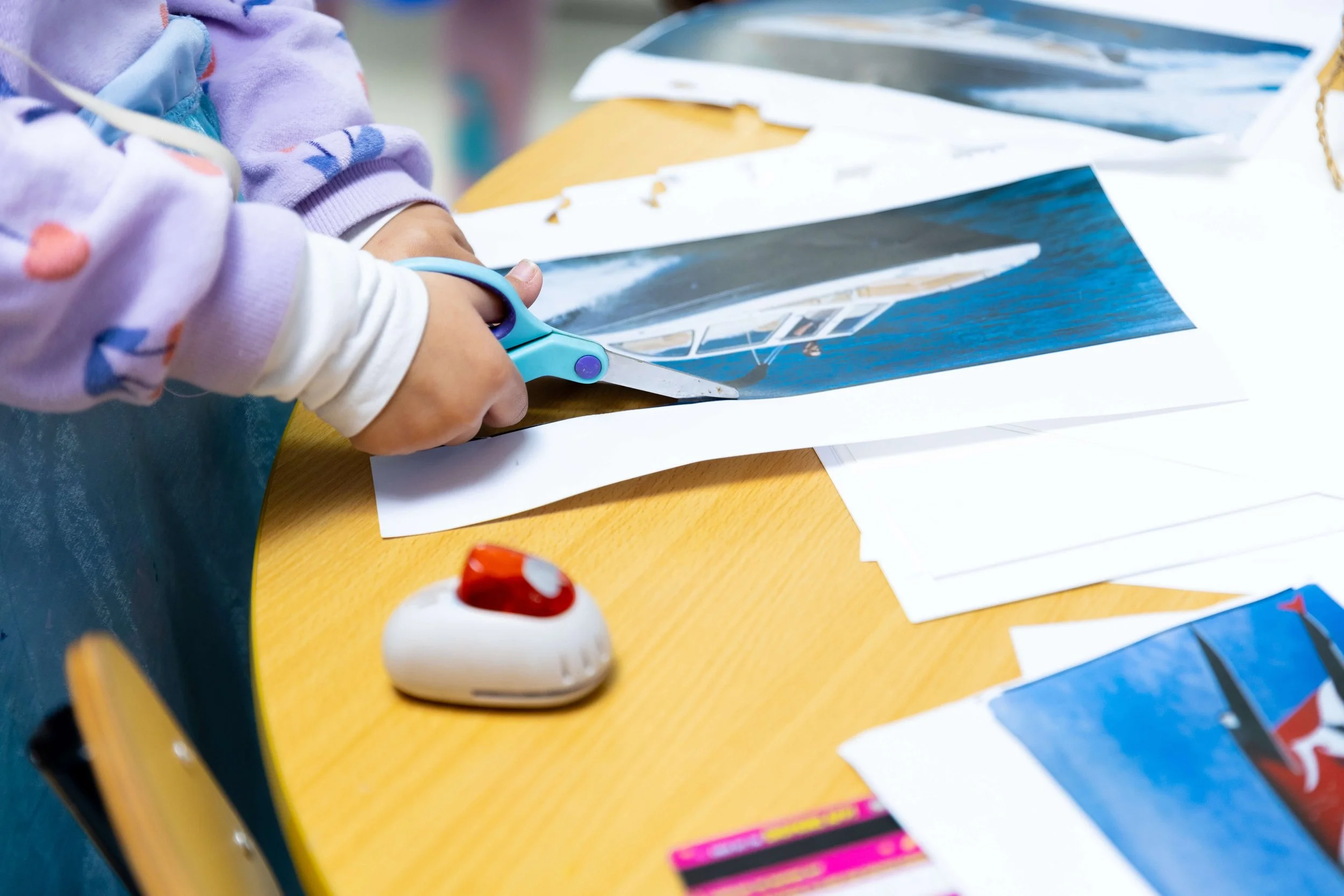 Child using scissors to cut a picture of a boat out of a sheet of paper on a table with stacks of papers and a small object resembling a white and red toy car nearby.