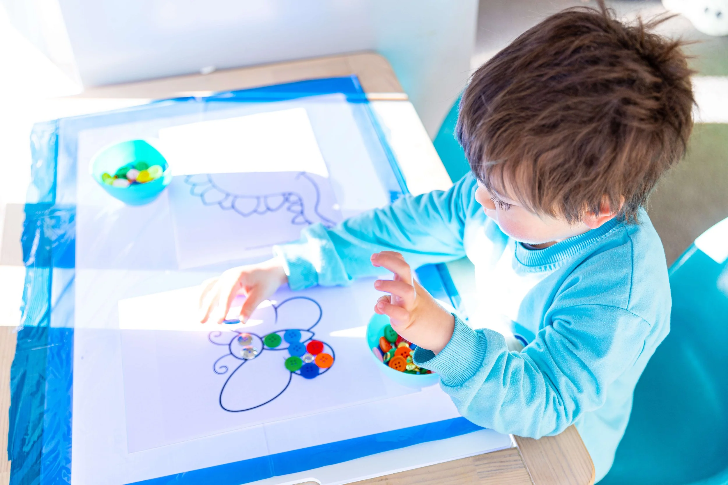 A young boy wearing a light blue long sleeve shirt is sitting at a table, decorating a butterfly shape on a piece of paper with colorful buttons.