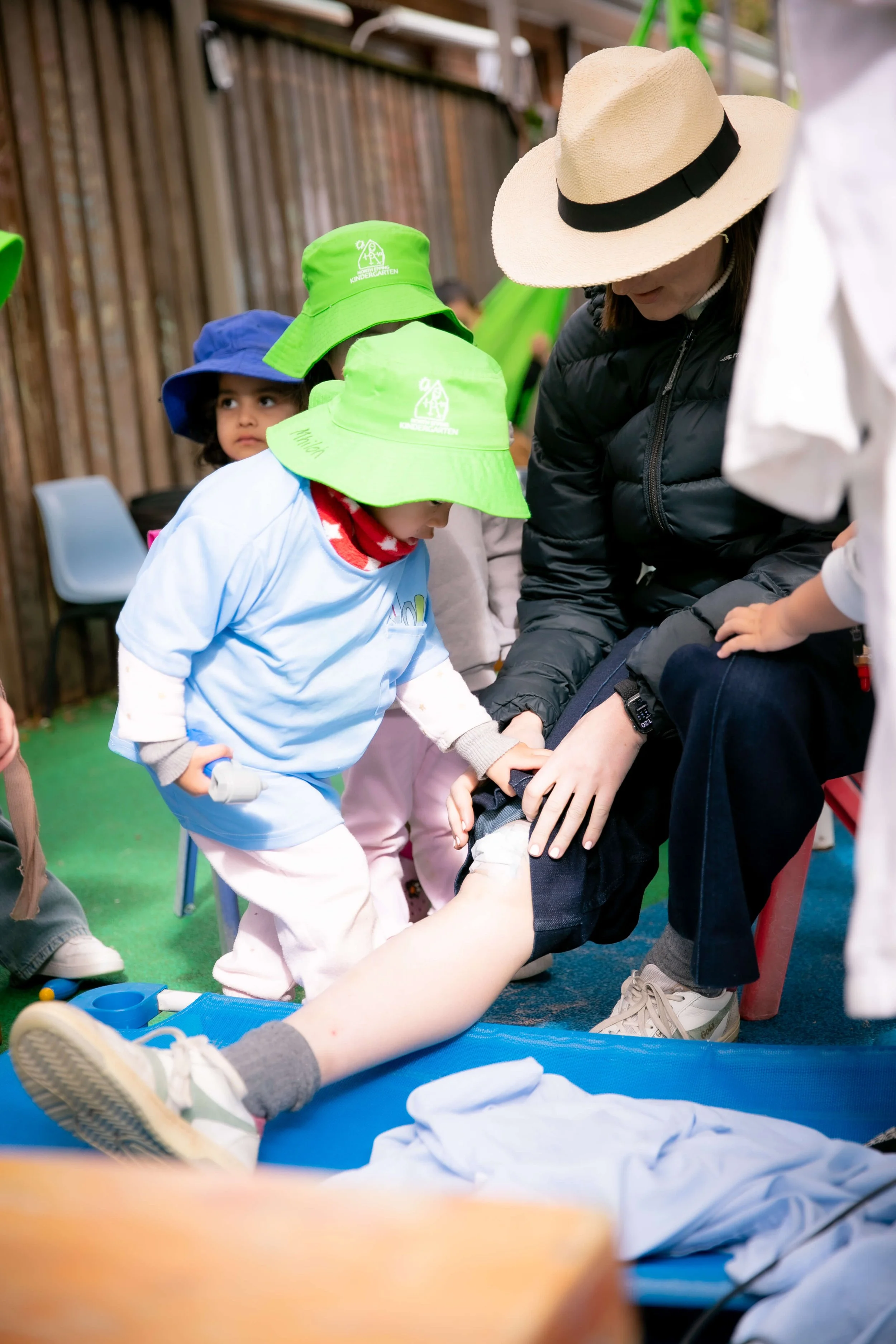 Children practicing first aid on a model leg, supervised by an instructor, in a classroom setting.