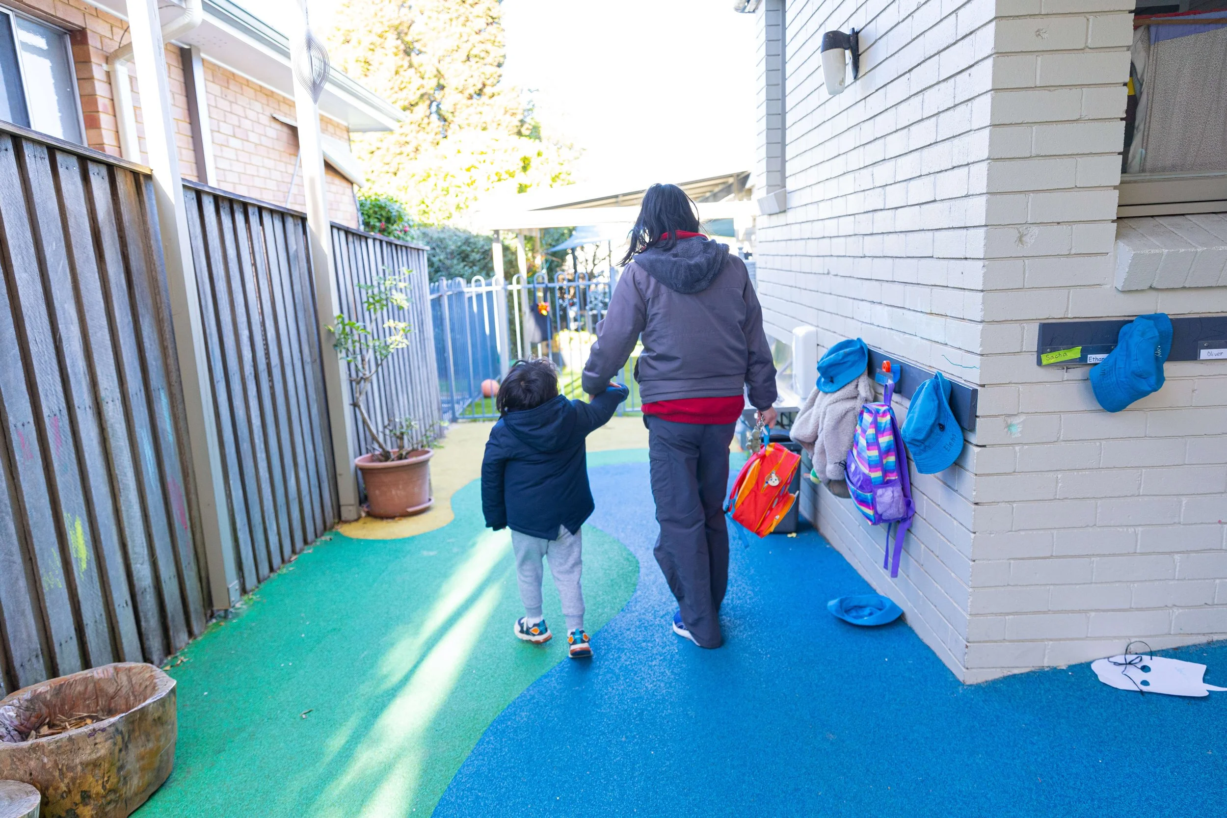 A woman and a young child walking on a colorful outdoor playground path, holding hands. The woman carries a backpack, and the child wears a dark jacket with white pants. There are outdoor play structures and a fence in the background.