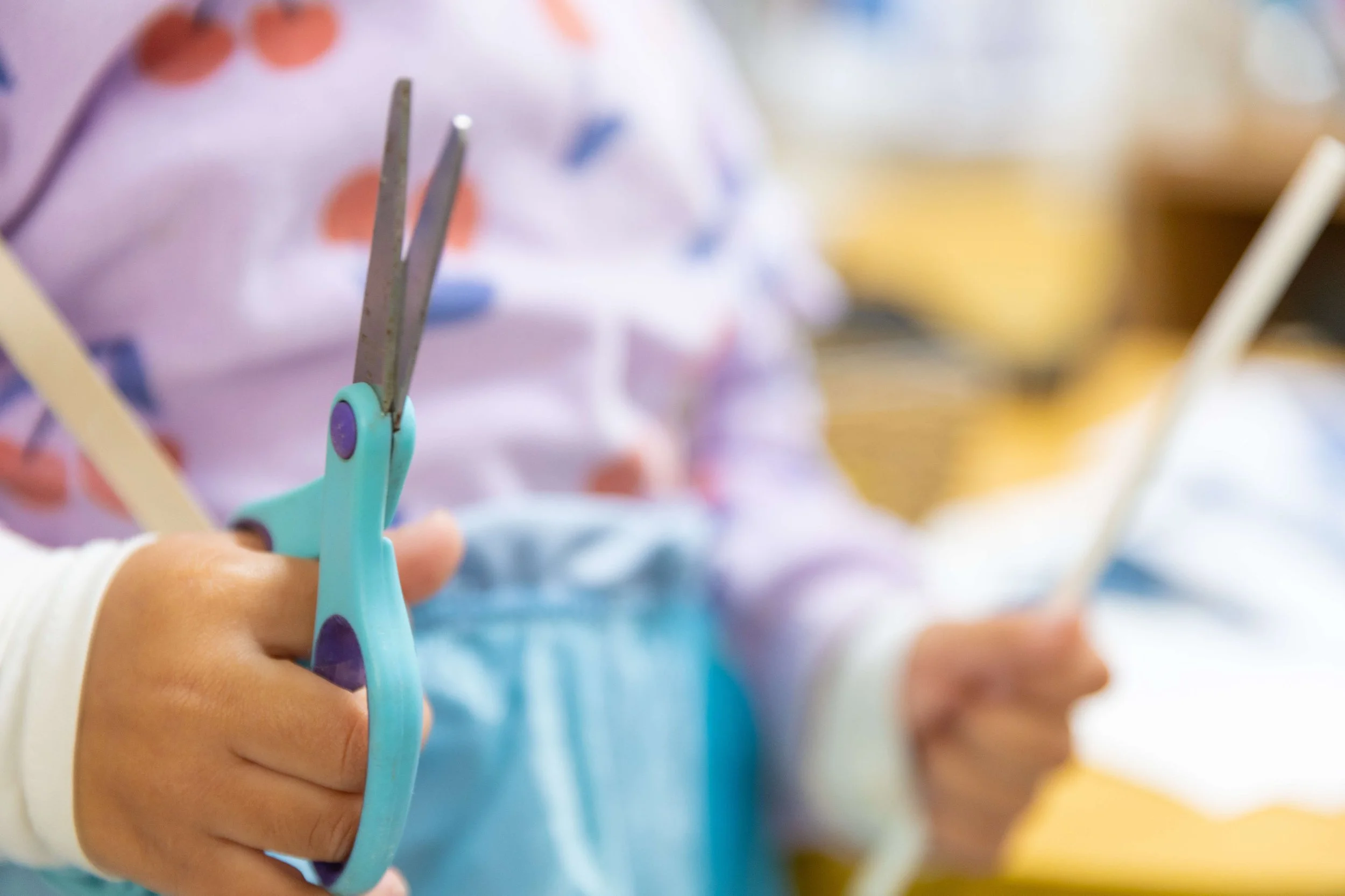 Close-up of a child's hand holding a piece of blue safety scissors, with a background showing a person holding a pair of chopsticks and wearing a colorful shirt.
