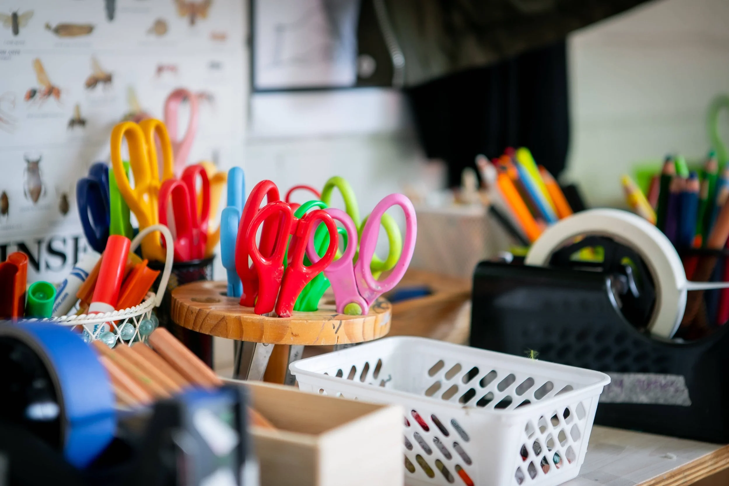A variety of colorful scissors, markers, and office supplies arranged on a cluttered desk or craft table.