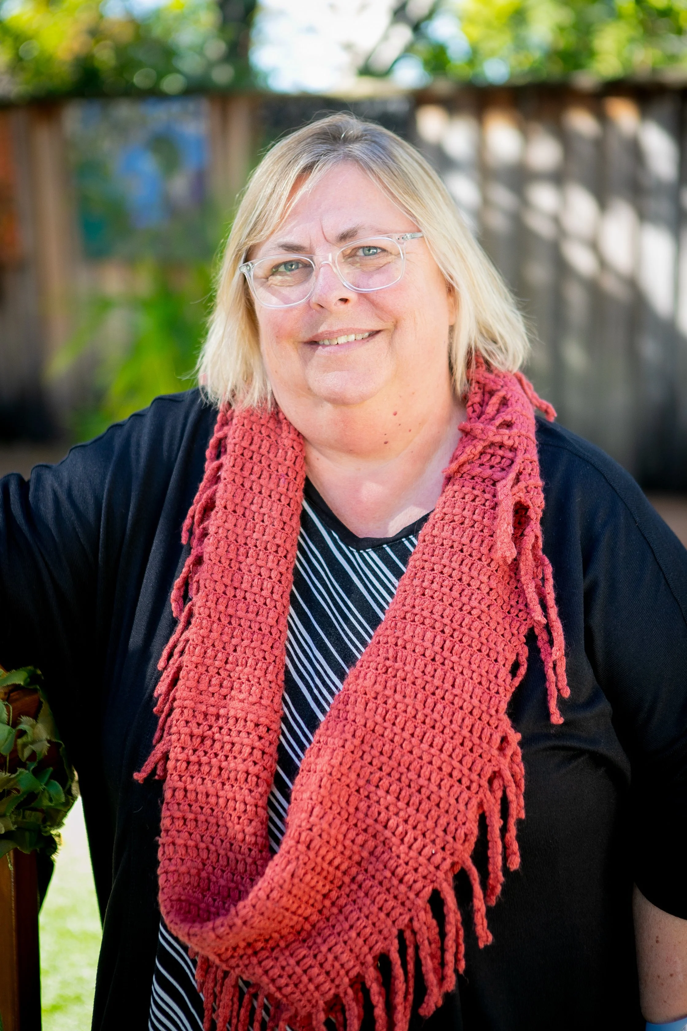 A woman with glasses and shoulder-length blonde hair wearing a black top, striped shirt, and a pink knitted scarf, standing outdoors in front of a blurred background of trees and a wooden fence, smiling at the camera.