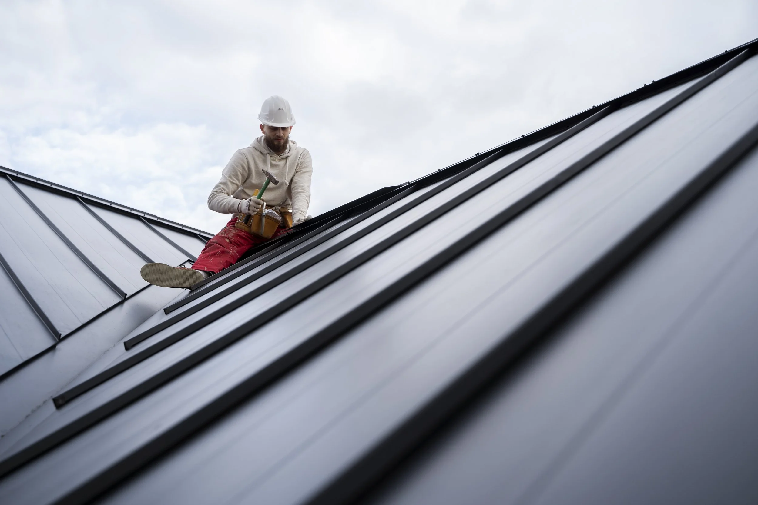 A man wearing a hard hat and work gloves is installing or repairing a metal roof, kneeling on the sloped roof with a hammer in hand, against a cloudy sky background.
