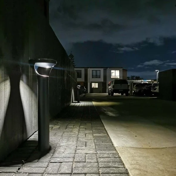 Nighttime view of a residential parking lot with cars, illuminated by outdoor lamps along a paved pathway and a sidewalk, with buildings in the background and a dark cloudy sky.