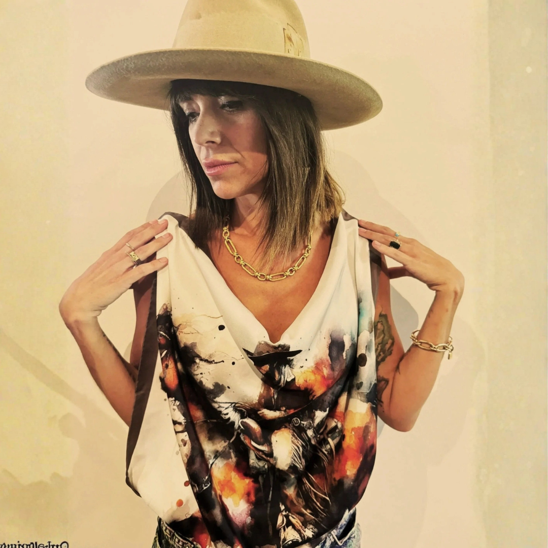 A woman wearing a large beige hat and a colorful sleeveless top, accessorized with gold jewelry, standing against a plain beige wall.