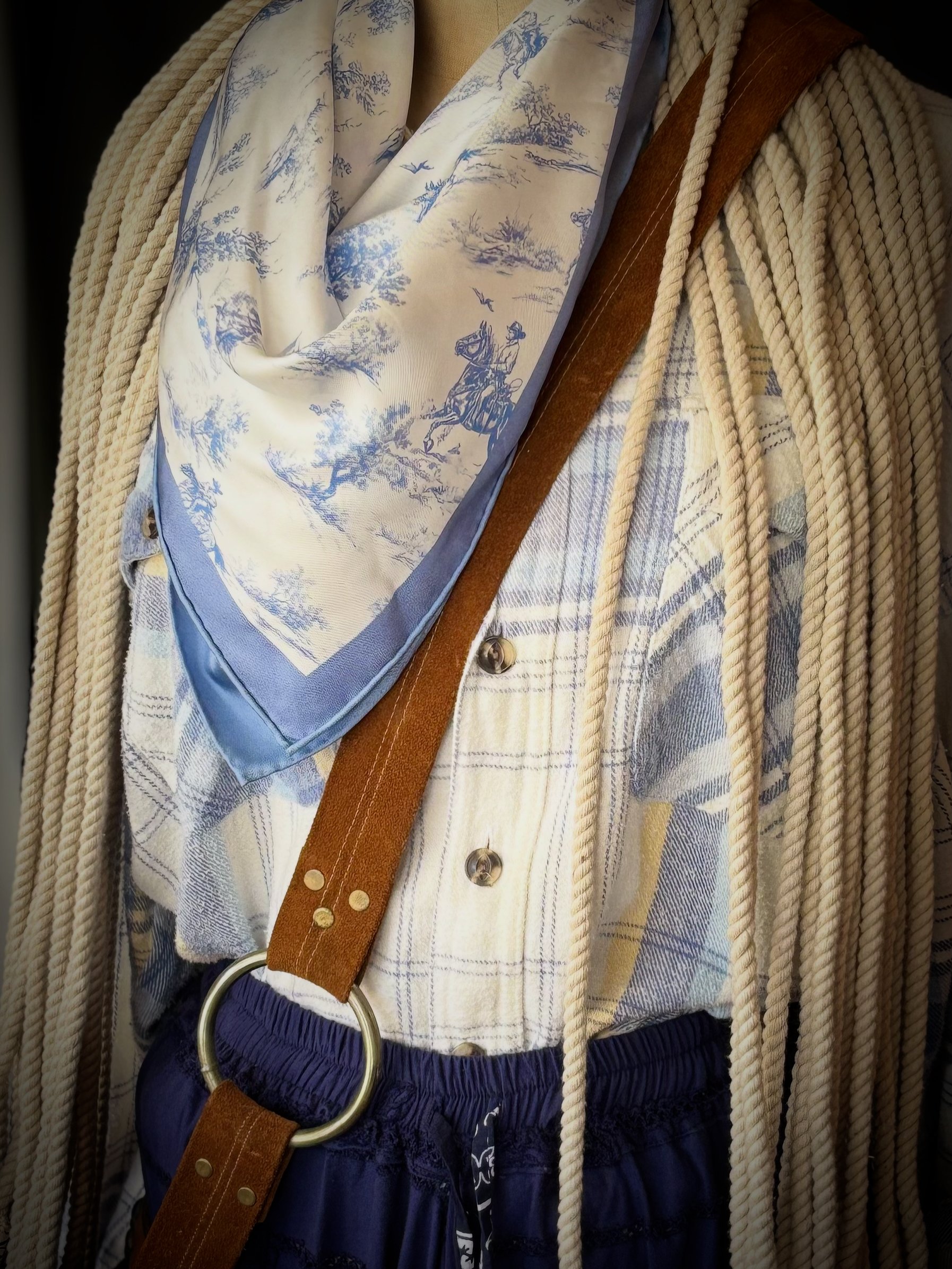 Close-up of a shirt with a plaid pattern, layered under a fringed shawl, and accessorized with a brown leather strap with a silver ring, a blue silk scarf with a pastoral scene, and navy shorts with a drawstring.
