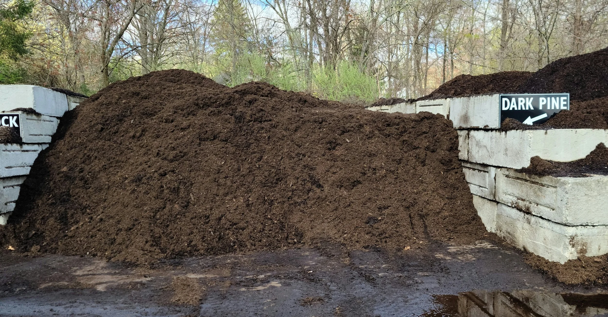 Large pile of dark soil in front of white brick walls with labeled sign that says 'Dark Pine' and partially visible sign pointing to 'Black.'