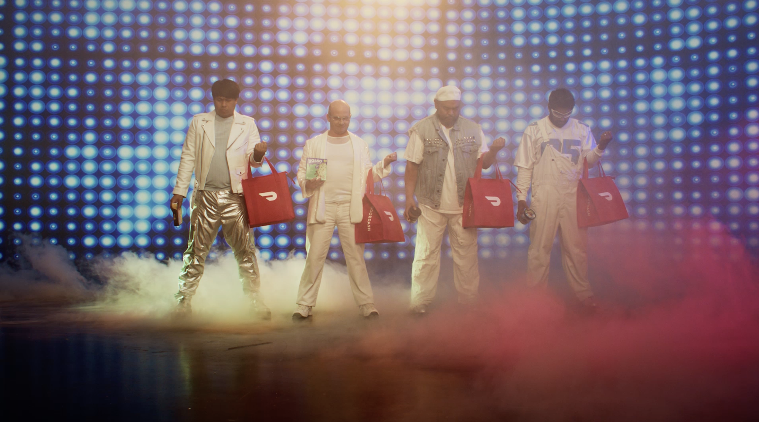 Five people standing on a stage with colorful lights and smoke, each holding a red shopping bag and some holding drinks, dressed in casual and shiny outfits.