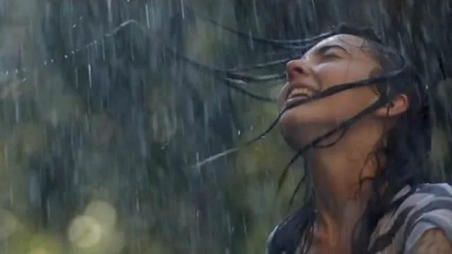 A woman standing in the rain with her head tilted back and eyes closed, water streaming down her face.