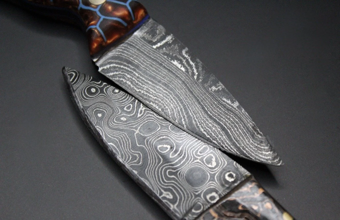 Close-up of a Damascus steel knife with a patterned blade and a decorative handle, placed on a dark surface.