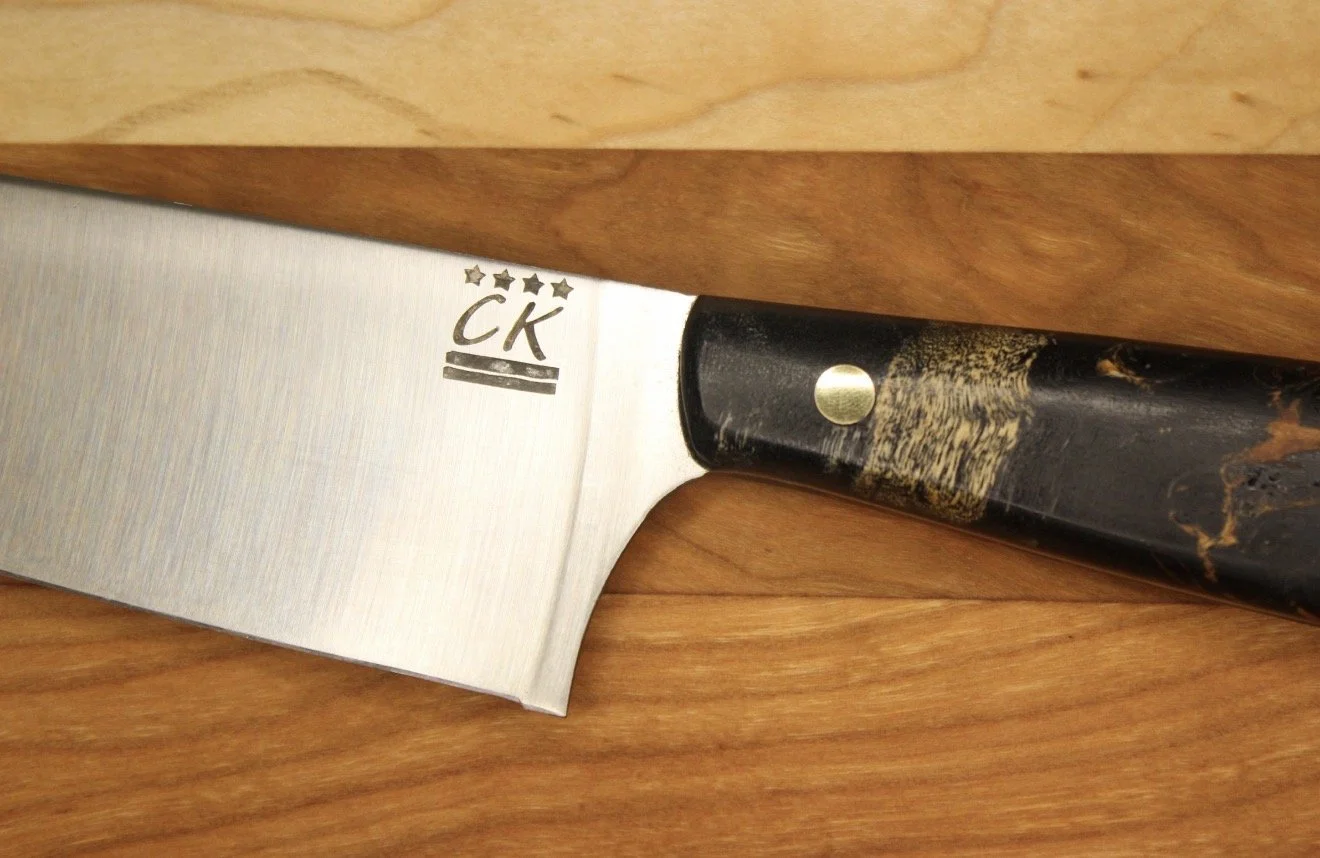 Close-up of a kitchen chef's knife with a stainless steel blade and a dark, handle, resting on a wooden surface.