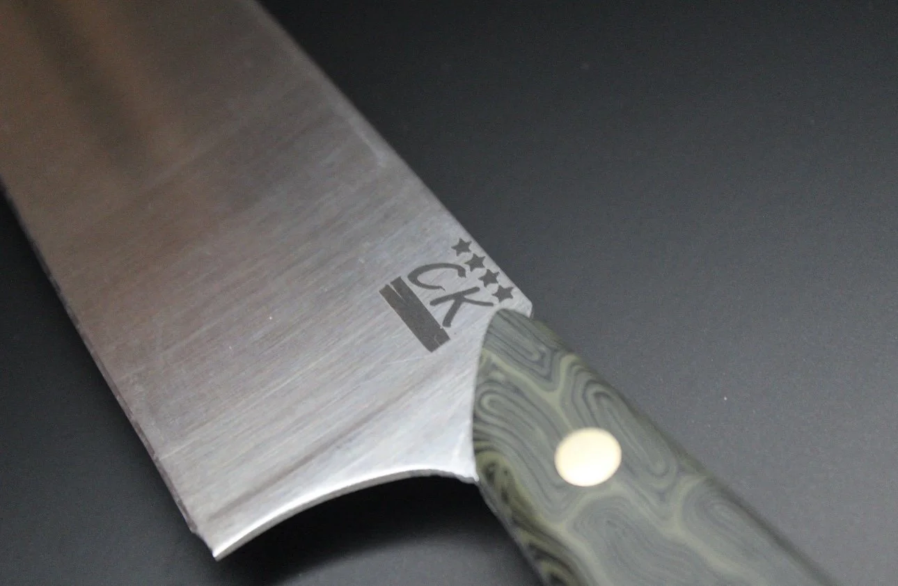 Close-up of a chef's knife with a patterned handle and a logo on the blade, with a dark background.