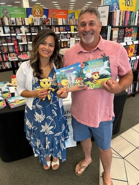 A woman and a man smiling and holding children's books inside a bookstore. The woman is wearing a blue floral dress and the man is wearing a pink shirt and shorts. Bookshelves and signs are visible in the background.