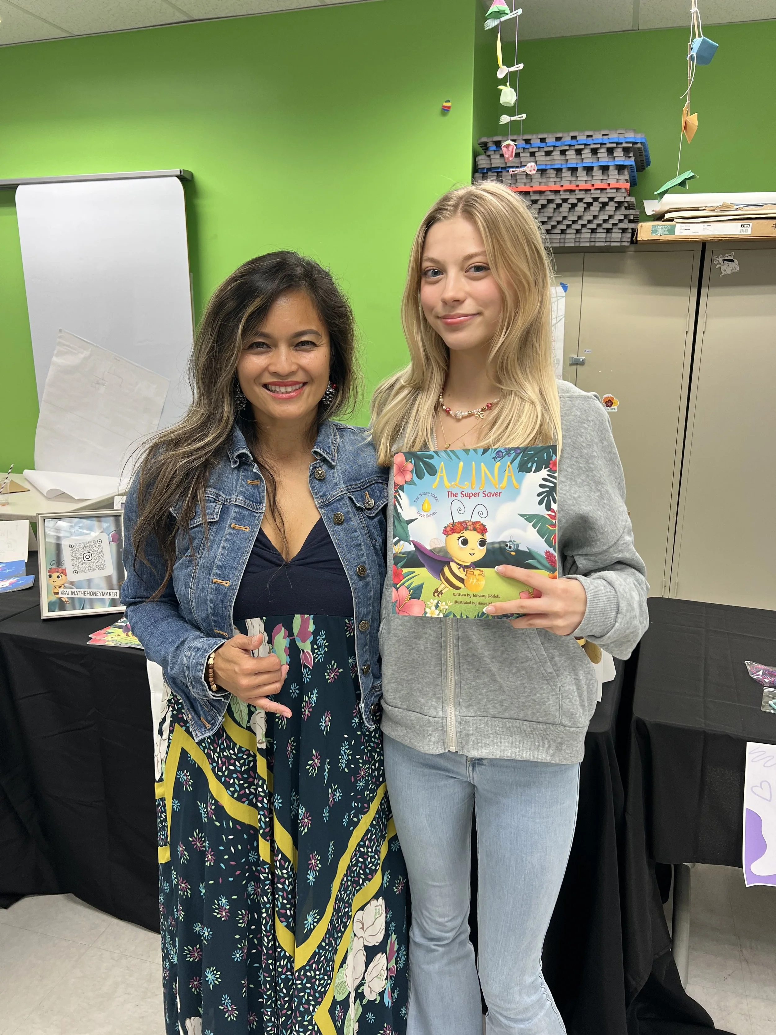 Two women standing together inside a room with green walls, holding a colorful children's book titled 'ALINA The Super Saver'. One woman has long dark hair, wearing a denim jacket, and the other has long blonde hair, wearing a gray hoodie. They are s