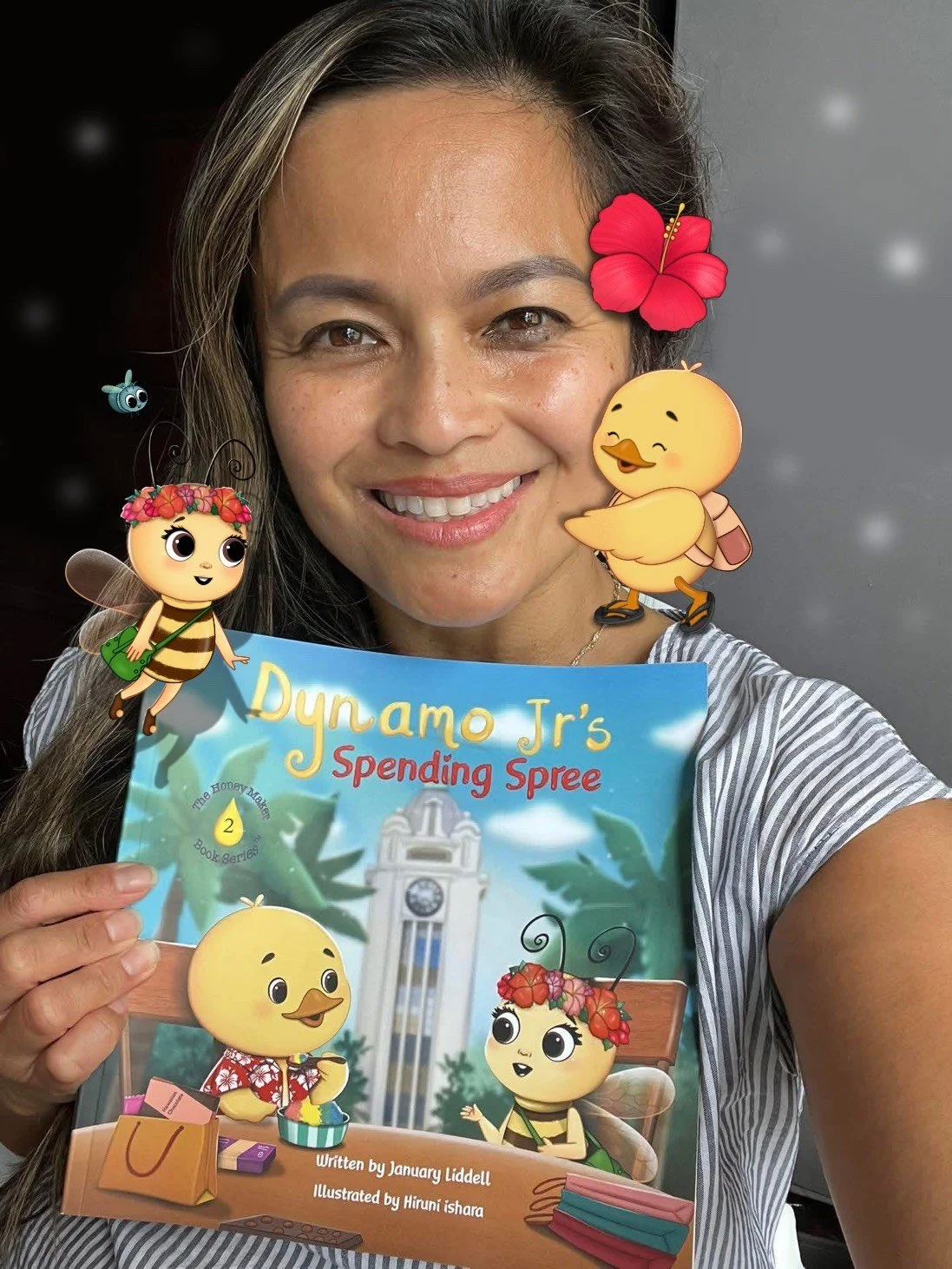 A woman with long hair smiling, holding a children's book titled 'Dynamo Jr's Spending Spree,' decorated with animated bee, chick, and flower characters.