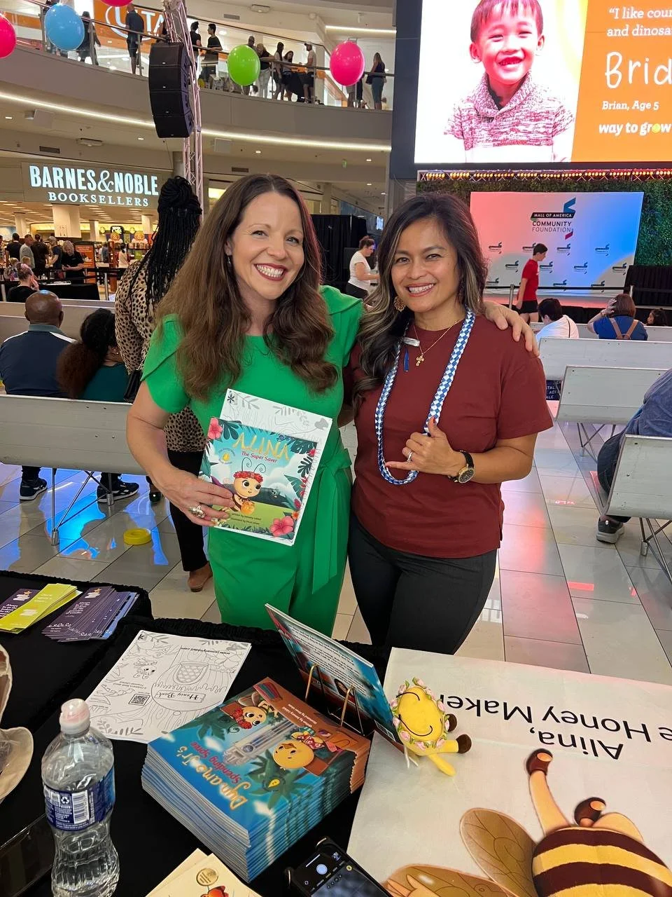 Two smiling women standing together at a book signing event inside a shopping mall, with a table in front of them displaying brochures and plush toys, and a large digital screen and a bookstore in the background.