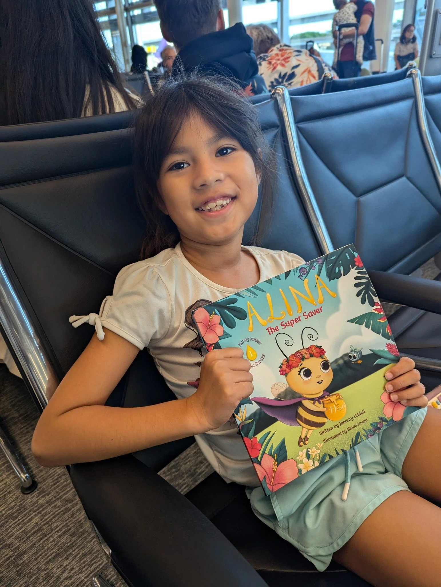 A young girl sitting in an airport holding a colorful children's book titled 'Alina The Super Saver' with a cheerful expression, surrounded by other travelers and airport seating.