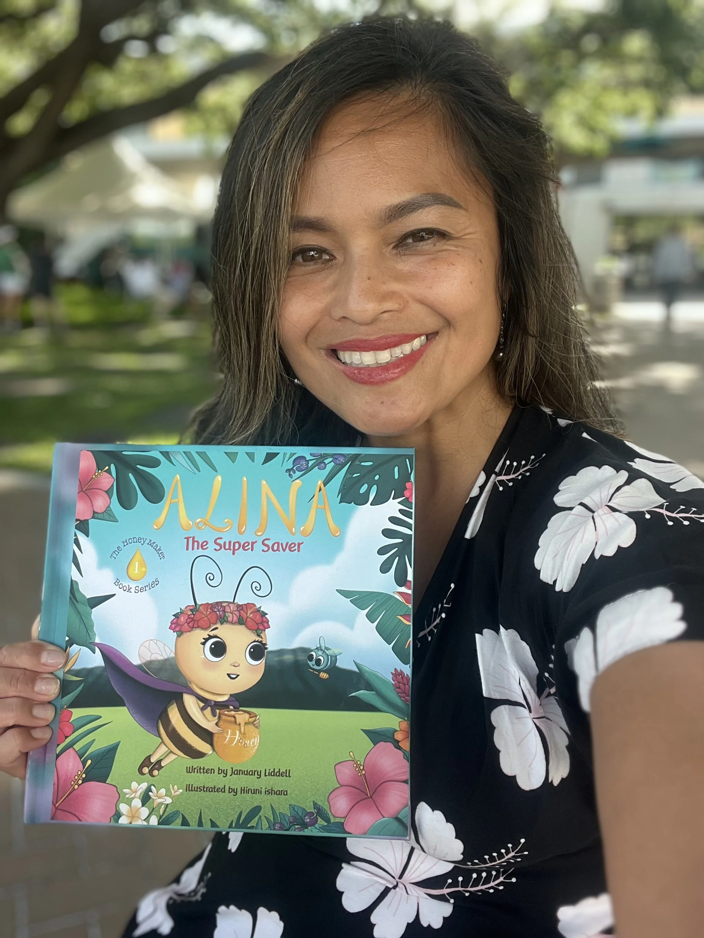 A woman is smiling and taking a selfie outdoors while holding a children's book titled 'Alina The Super Saver.' The book cover depicts a cartoon bee character with a purple cape, holding a honey pot, surrounded by colorful flowers and greenery. The w