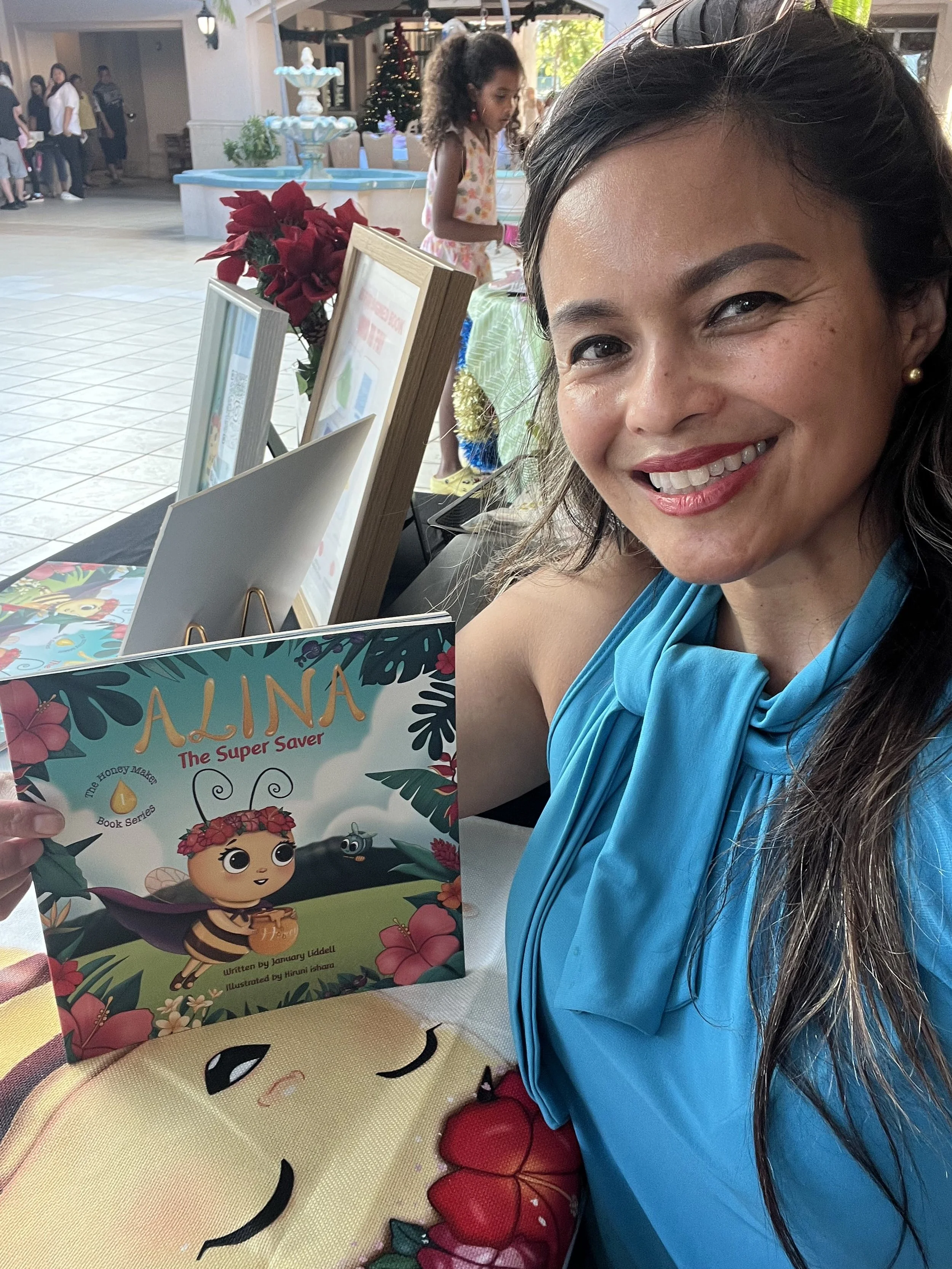 A woman in a blue dress holding a children's book titled 'Alina The Super Saver' with a smiling girl in a pink dress in the background at an indoor event.