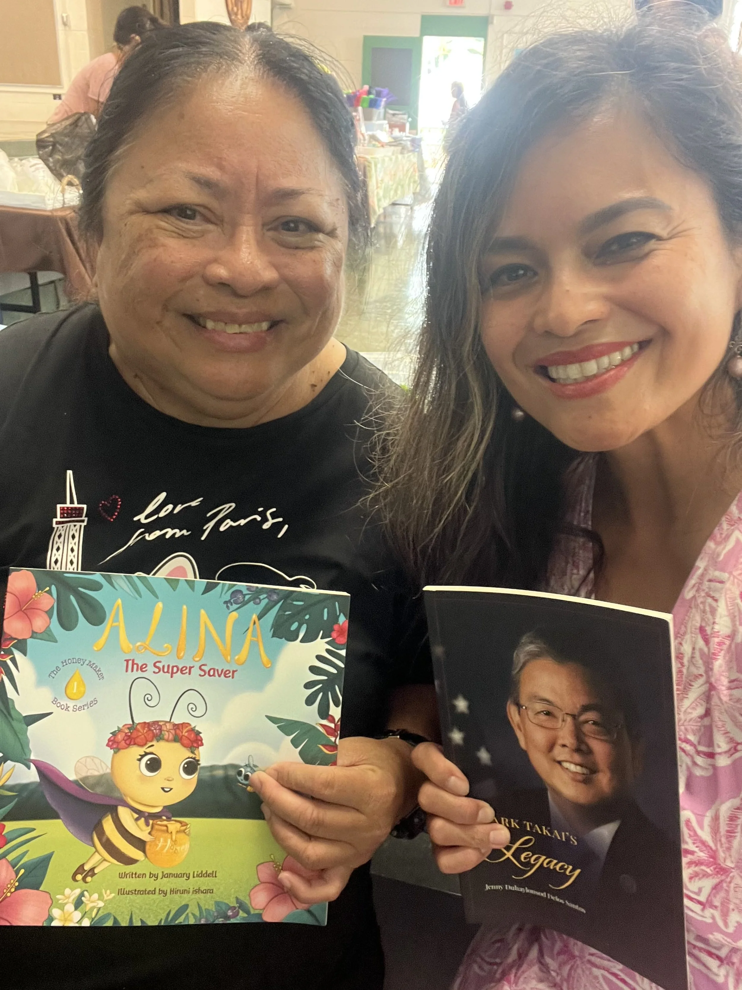 Two women smiling and holding books, one is holding a colorful children's book titled 'ALINA The Super Saver' and the other is holding a black-and-white portrait-oriented book titled 'ARK TAKAT'S Legacy', in an indoor setting with other people and ta