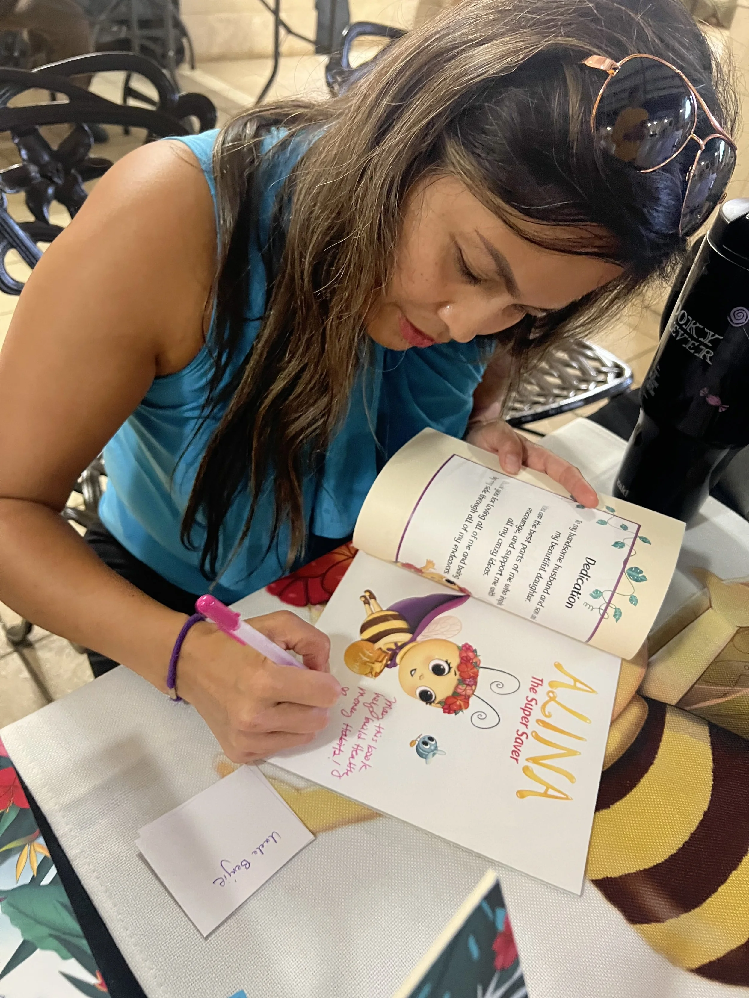 A woman with long brunette hair, wearing sunglasses on her head and a blue sleeveless top, is signing a book titled 'AALLINA The Super Saver' with a pink marker. The book features cartoon characters, including a bee and a small blue creature. She is 