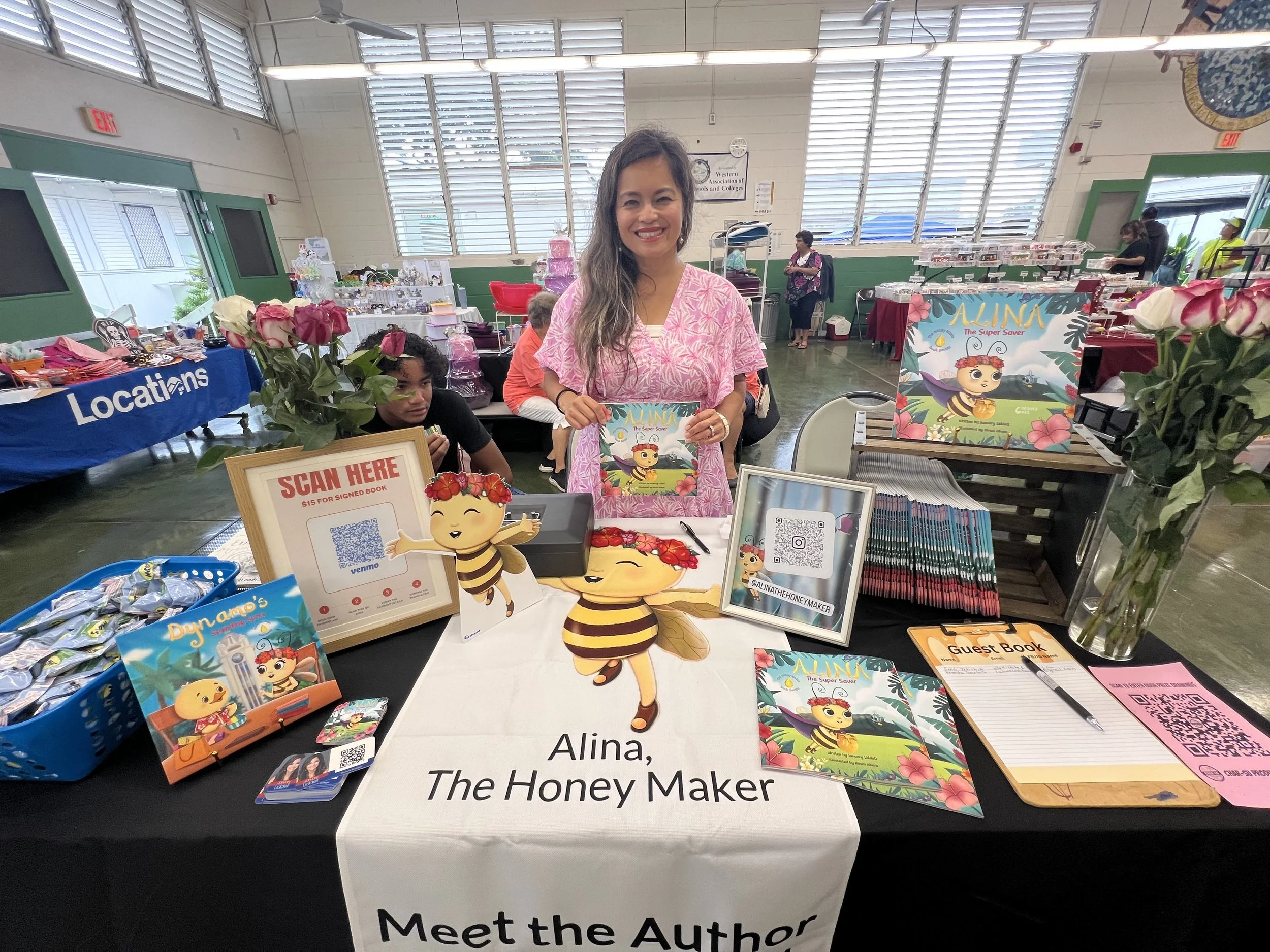 A woman at a booth named 'Alina, The Honey Maker' is smiling and holding a book titled 'Alina The Super Saver', surrounded by promotional materials and decorations. The booth is decorated with pink roses in vases, cartoon bee cutouts, and various mer