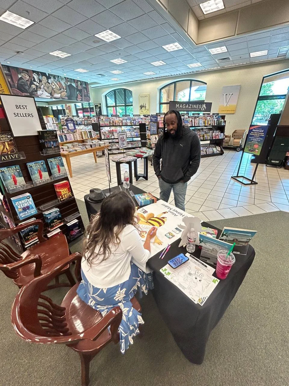 A young girl sits at a table signing a book for a smiling man in a bookstore. The table has a water bottle, a pink drink, and coloring pages. Bookshelves and posters are visible in the background.