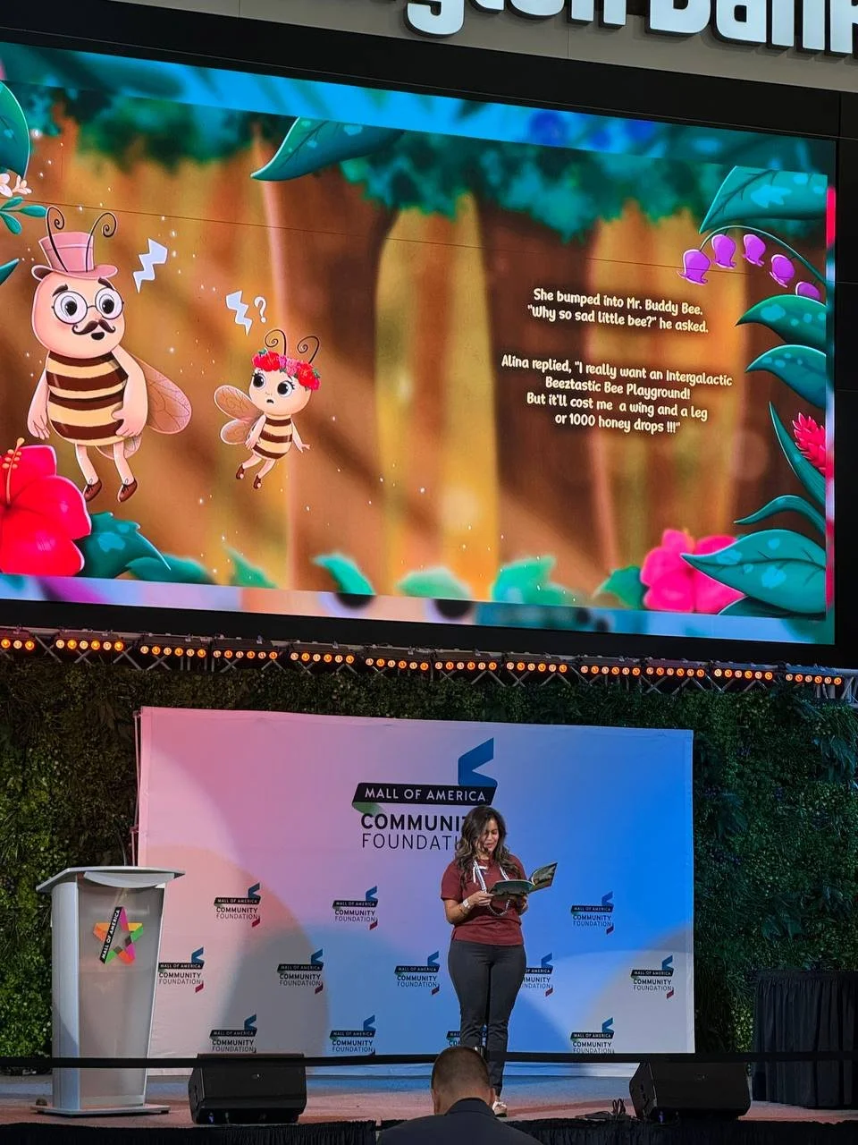 Large screen displaying animated cartoon bees with a butterfly and flowers in a forest scene; a dialogue between bees about a playground, along with a woman reading on stage at a community event.
