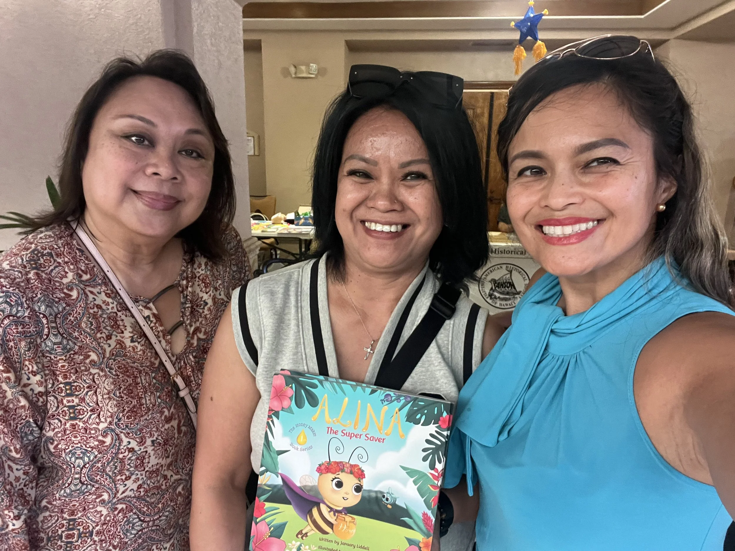 Three women smiling at a gathering, one holding a colorful children's book titled 'ALINA The Super Saver'. The background shows a table with various items and a sign from an organization.