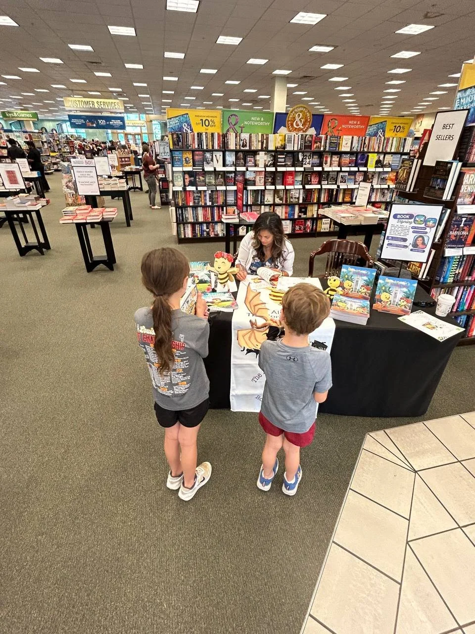 A woman signing books at a table inside a bookstore, with two children standing in front of her. The bookstore has shelves filled with books and signs promoting discounts and new releases.