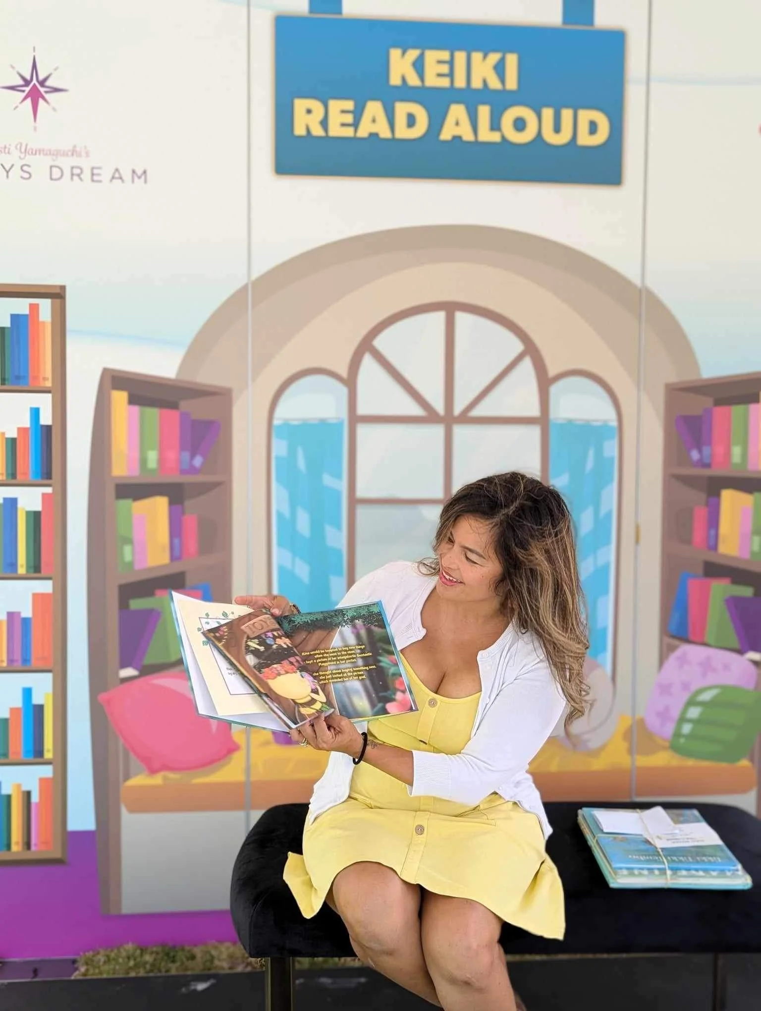 A woman sitting on a black bench reading a colorful children's book in front of a backdrop advertising a children's reading program called 'Keiki Read Aloud.' The backdrop depicts a cozy library scene with bookshelves and large windows.