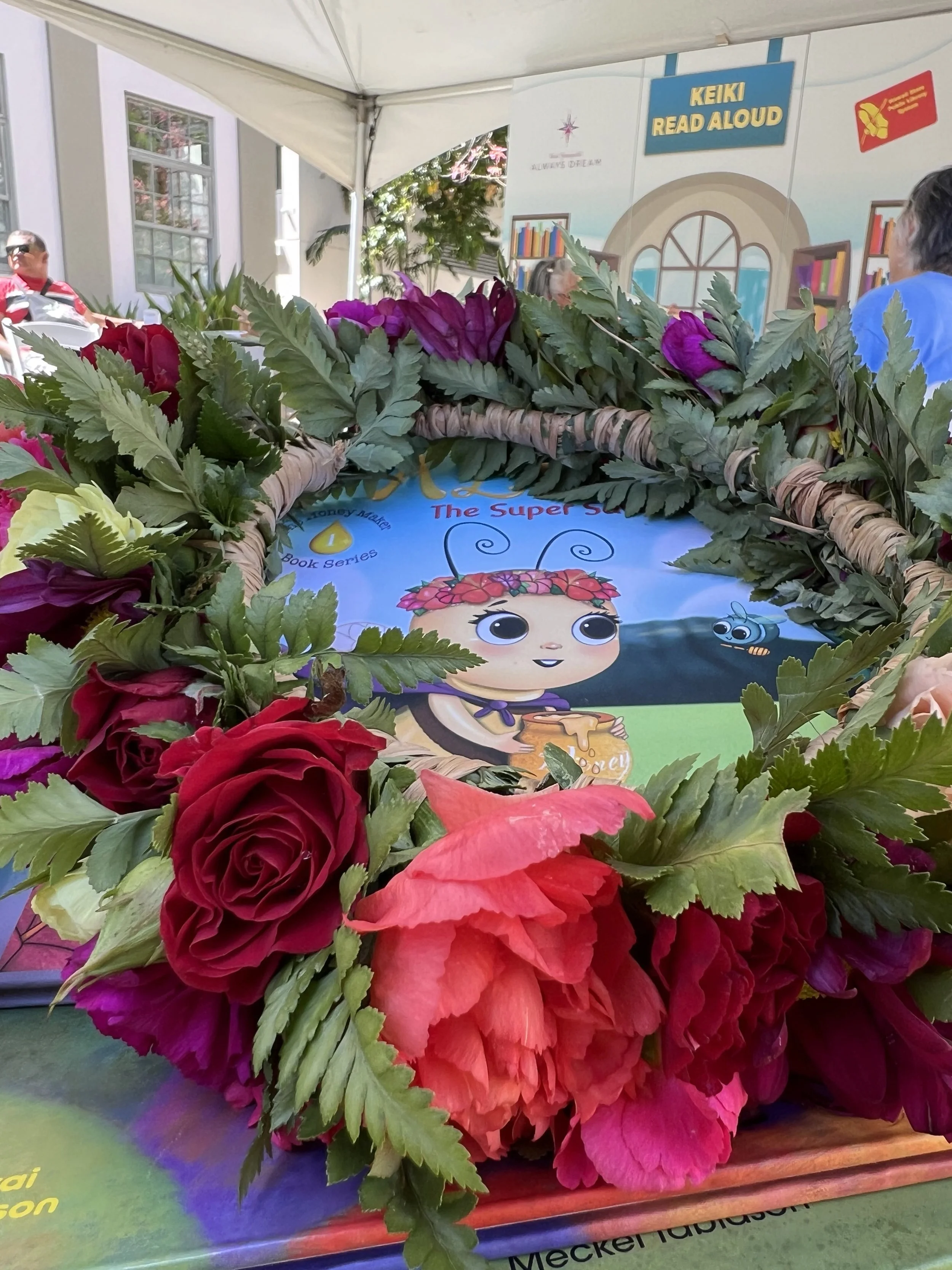 A book cover titled 'The Super Bee' surrounded by a floral wreath with roses and greenery, at a book event or display.